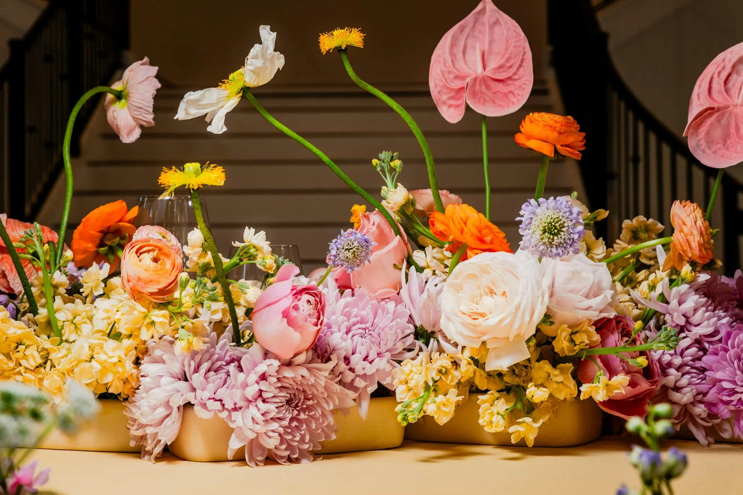 A colorful arrangement of various flowers, including pink roses, orange poppies, purple scabiosa, and white peonies, displayed in a yellow container with a blurred staircase in the background.