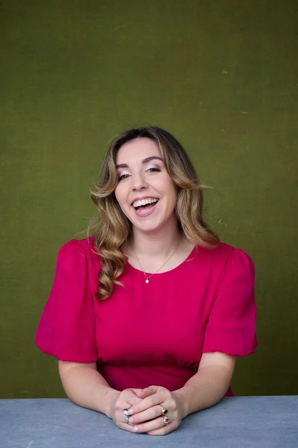 Woman with wavy blonde hair smiling, wearing a pink dress and rings, sitting at a table against a green background.