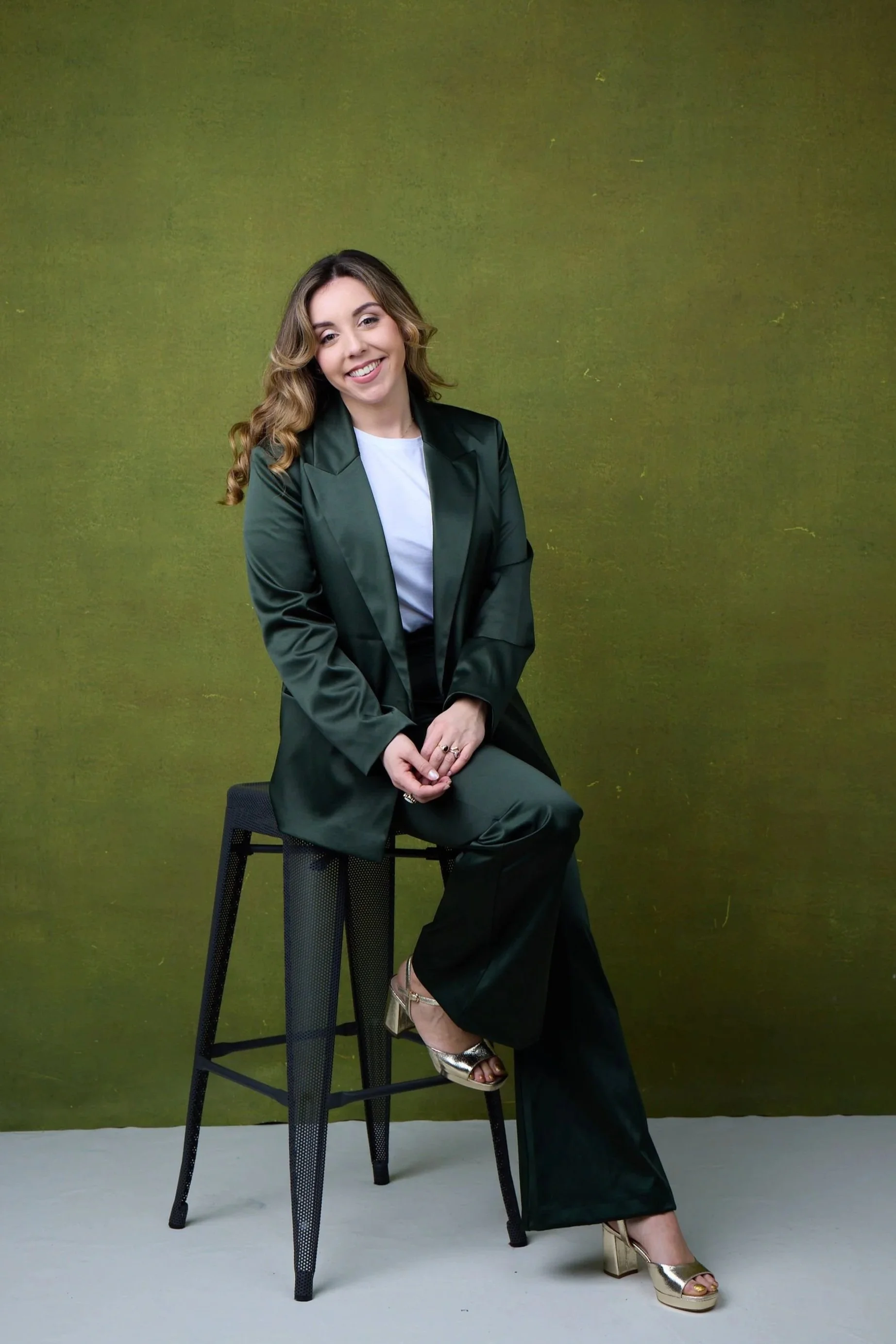 A woman sitting on a tall black stool against a textured green background, wearing a dark green blazer, white top, dark green wide-leg pants, and metallic gold high heels, smiling at the camera.