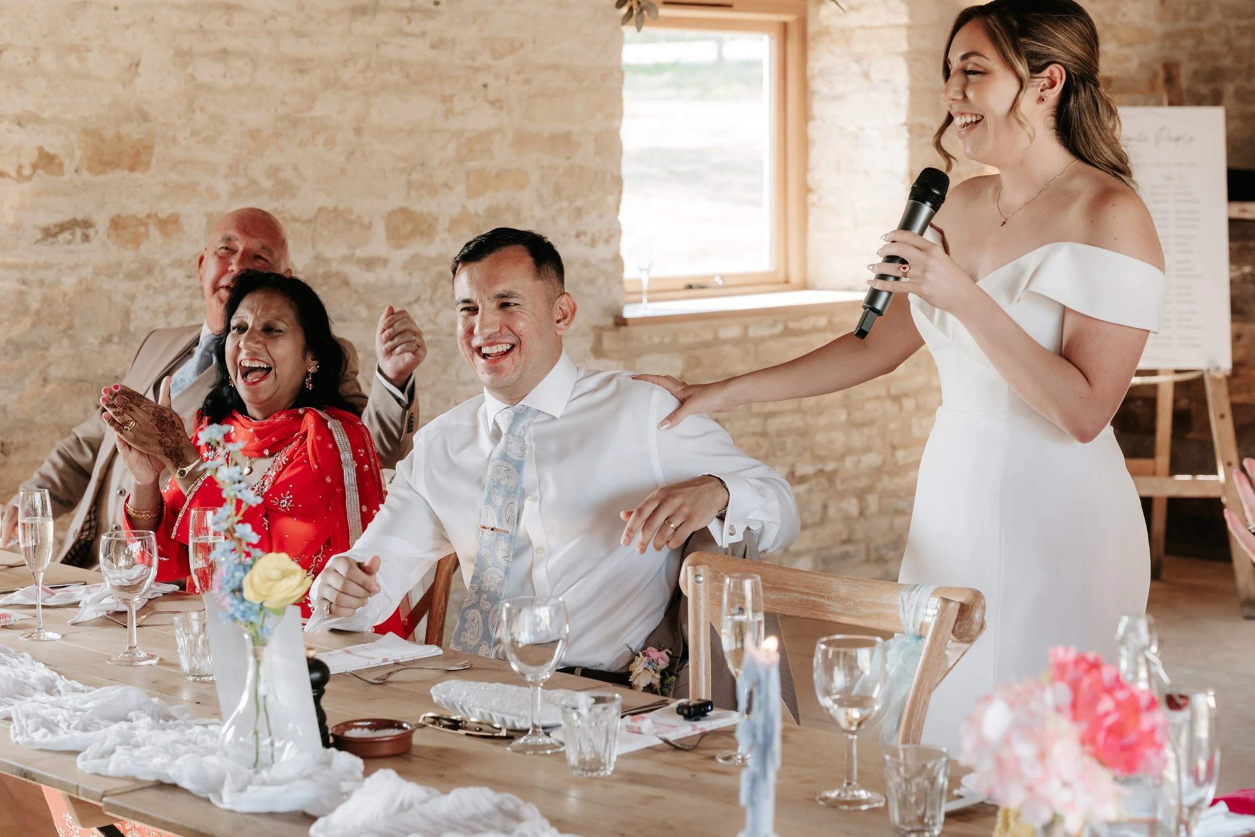 A woman in a white dress is giving a speech at a wedding reception, smiling as a man in a white shirt and tie laughs, with a woman in red and two older men also smiling and clapping in the background.