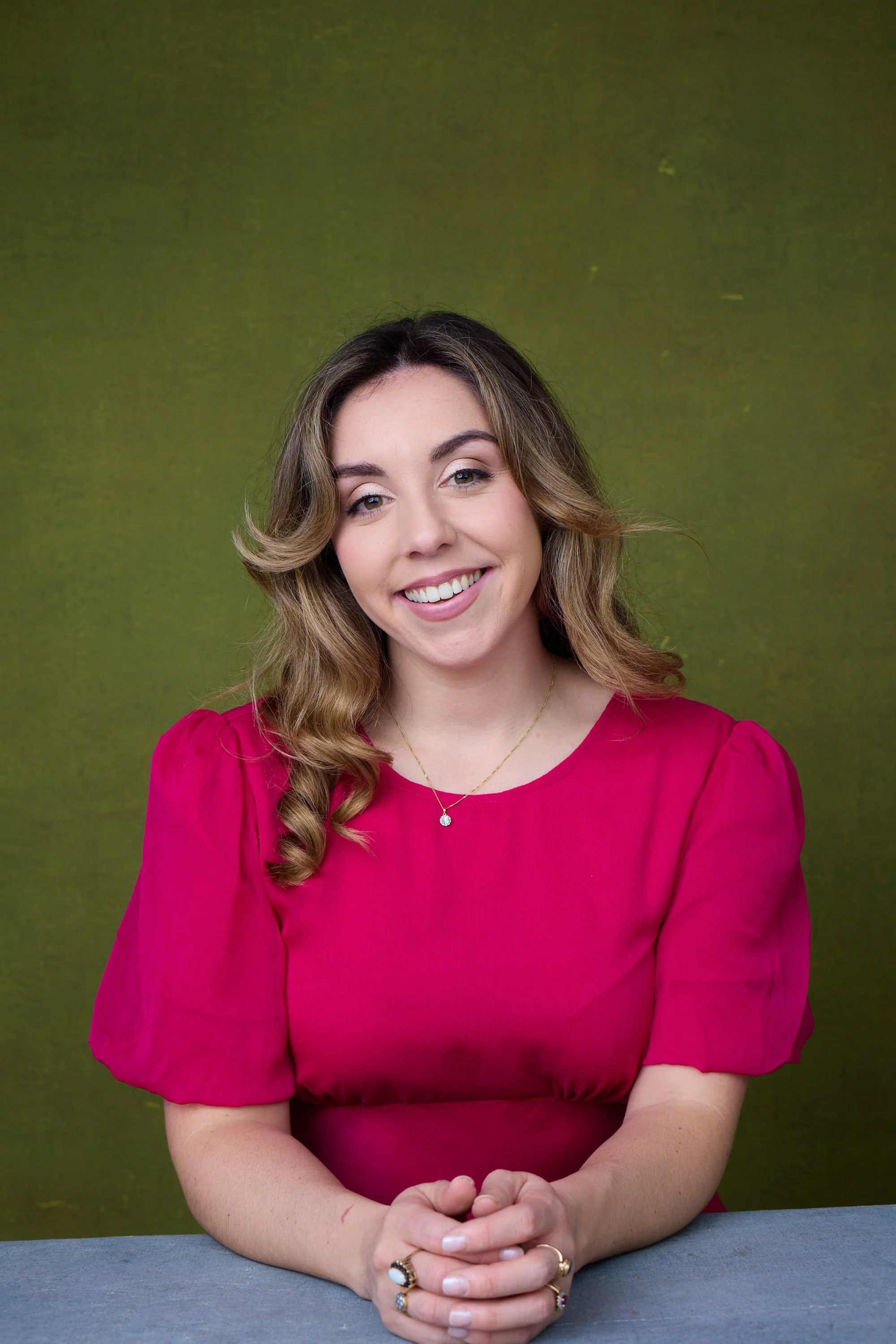 A woman with wavy brown hair, wearing a bright pink blouse with puffed sleeves, smiling sitting at a table with hands clasped, against a green textured background.