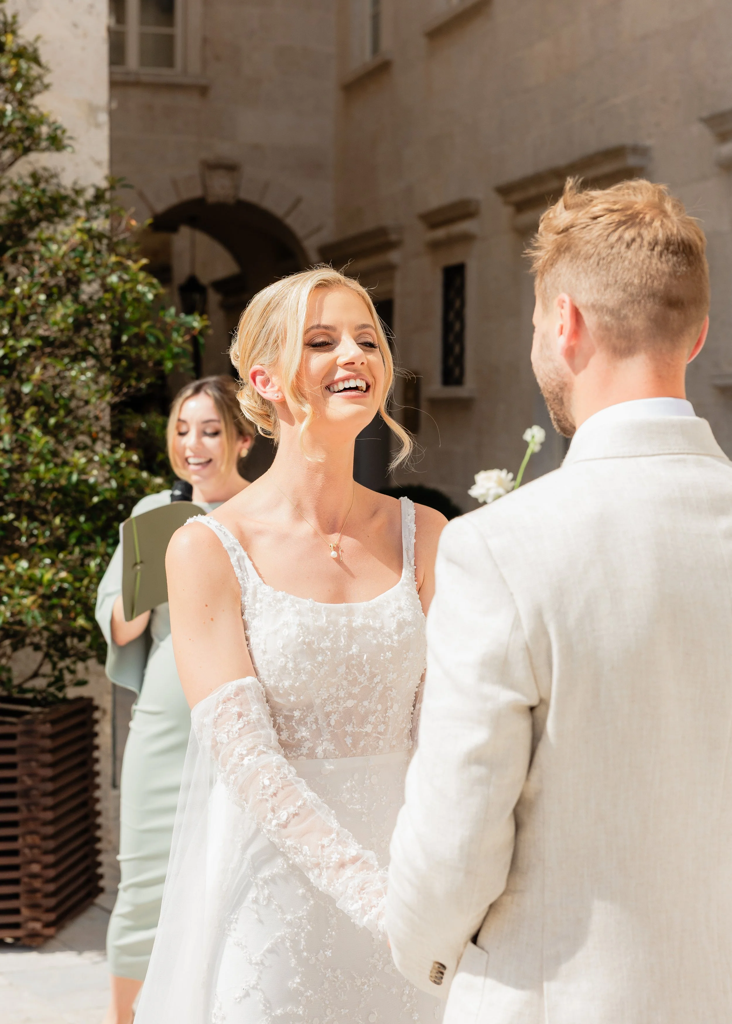 A smiling bride with blonde hair in a wedding dress holding hands with groom in a cream-colored suit during outdoor wedding ceremony.