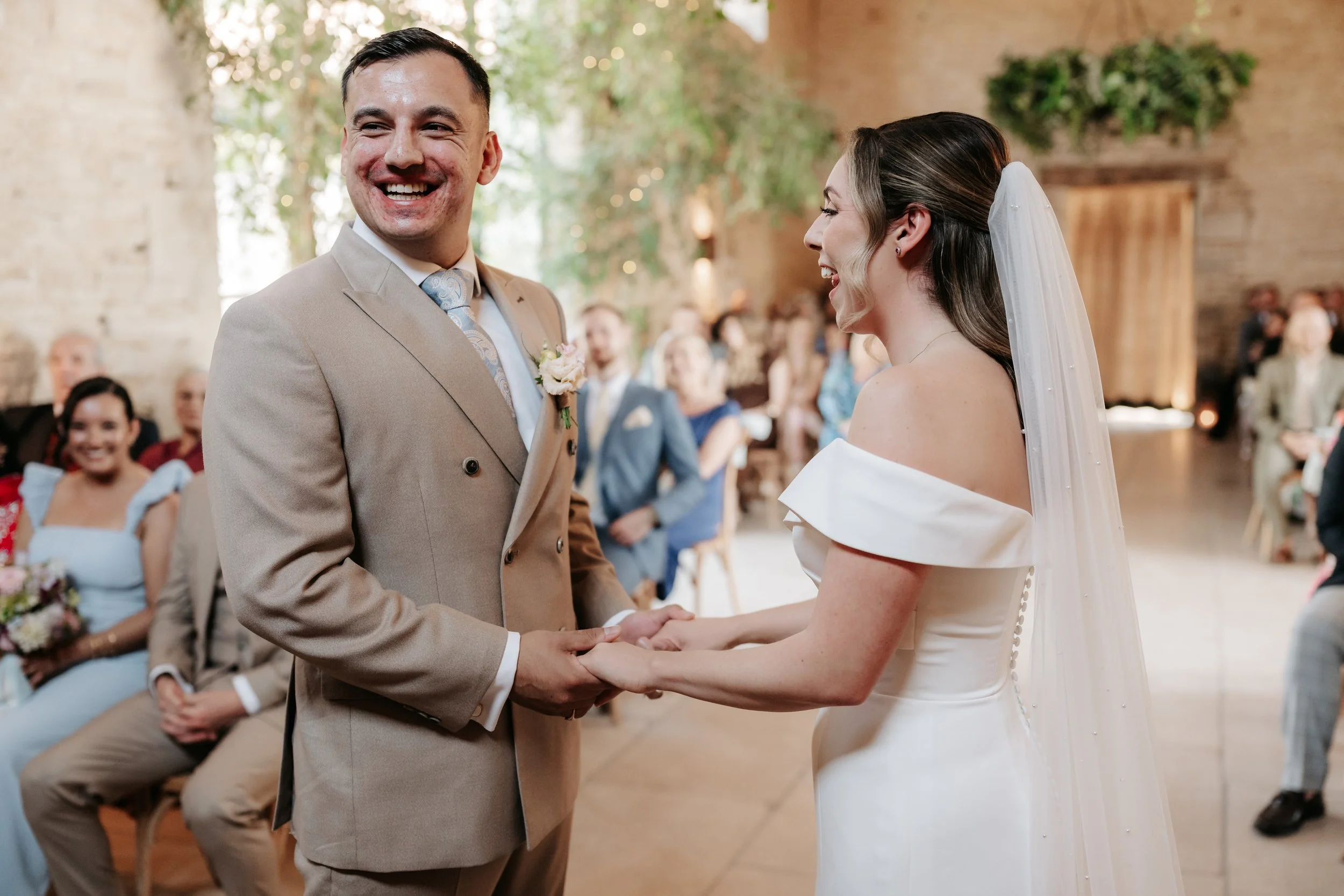A bride and groom holding hands and smiling during their wedding ceremony in a rustic venue with seated guests watching, some holding flowers.