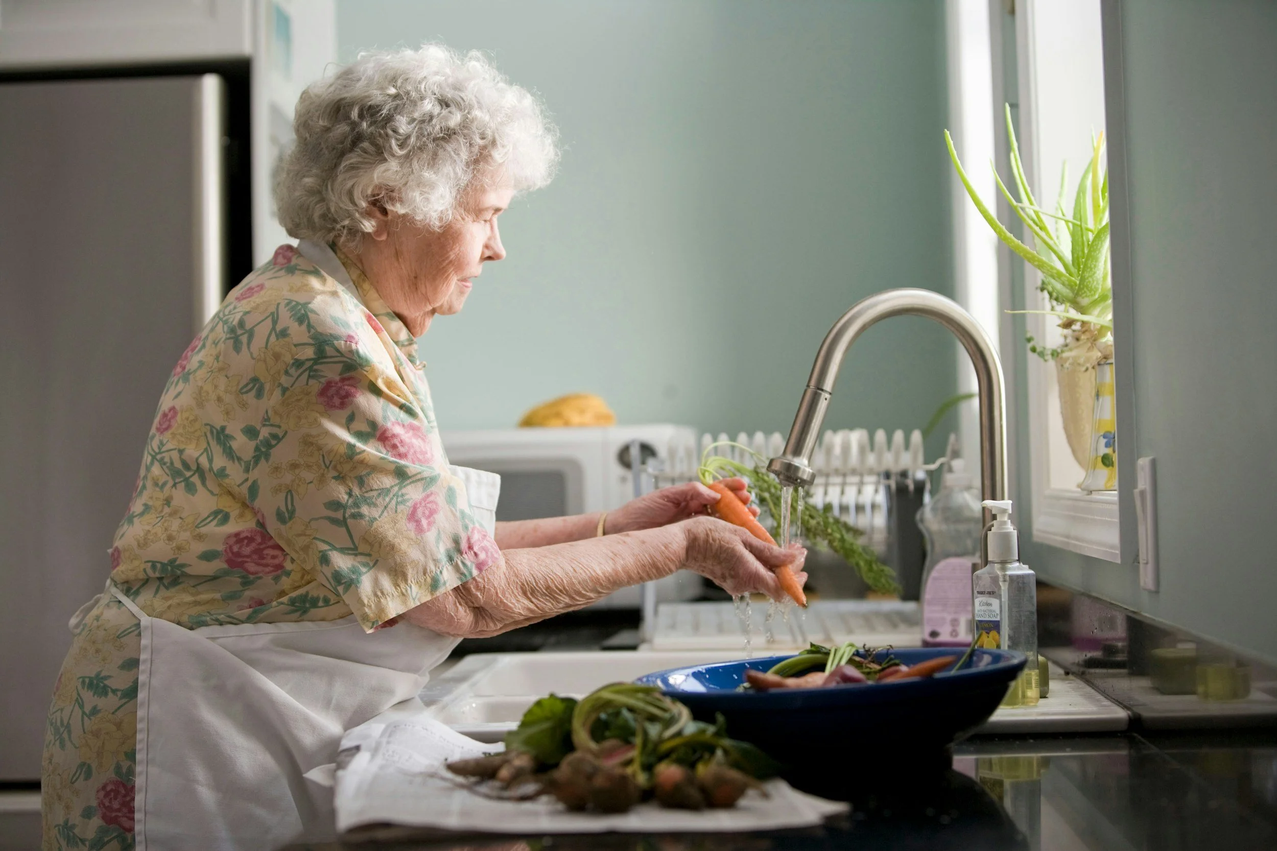 An elderly woman with curly gray hair washing carrots at a kitchen sink with a silver faucet, surrounded by fresh vegetables and houseplants, wearing a floral blouse and white apron.
