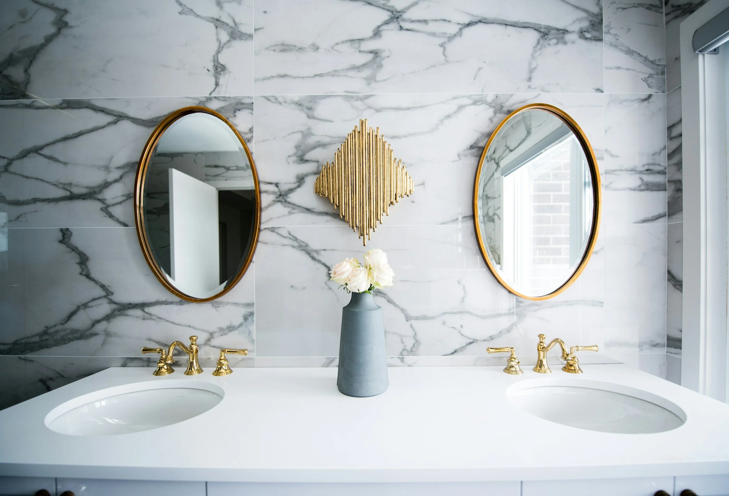 A bathroom vanity with two oval mirrors, a gray vase with white flowers, and gold fixtures, set against a marble wall.