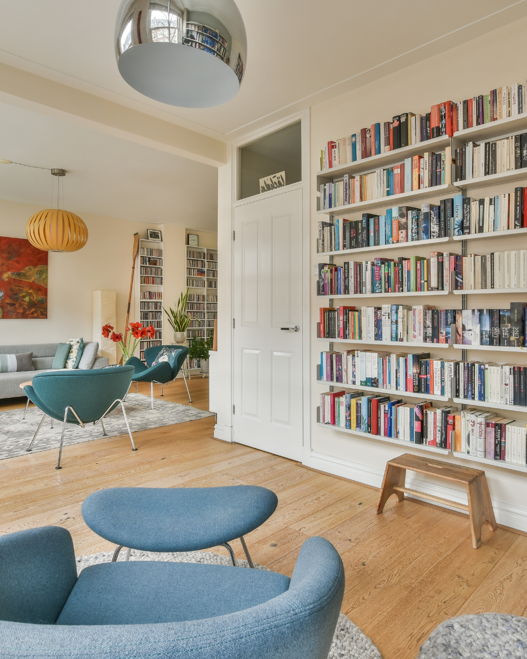 Living room with a large bookshelf filled with books, seating area with blue chairs, a gray sofa, plants, and artwork.