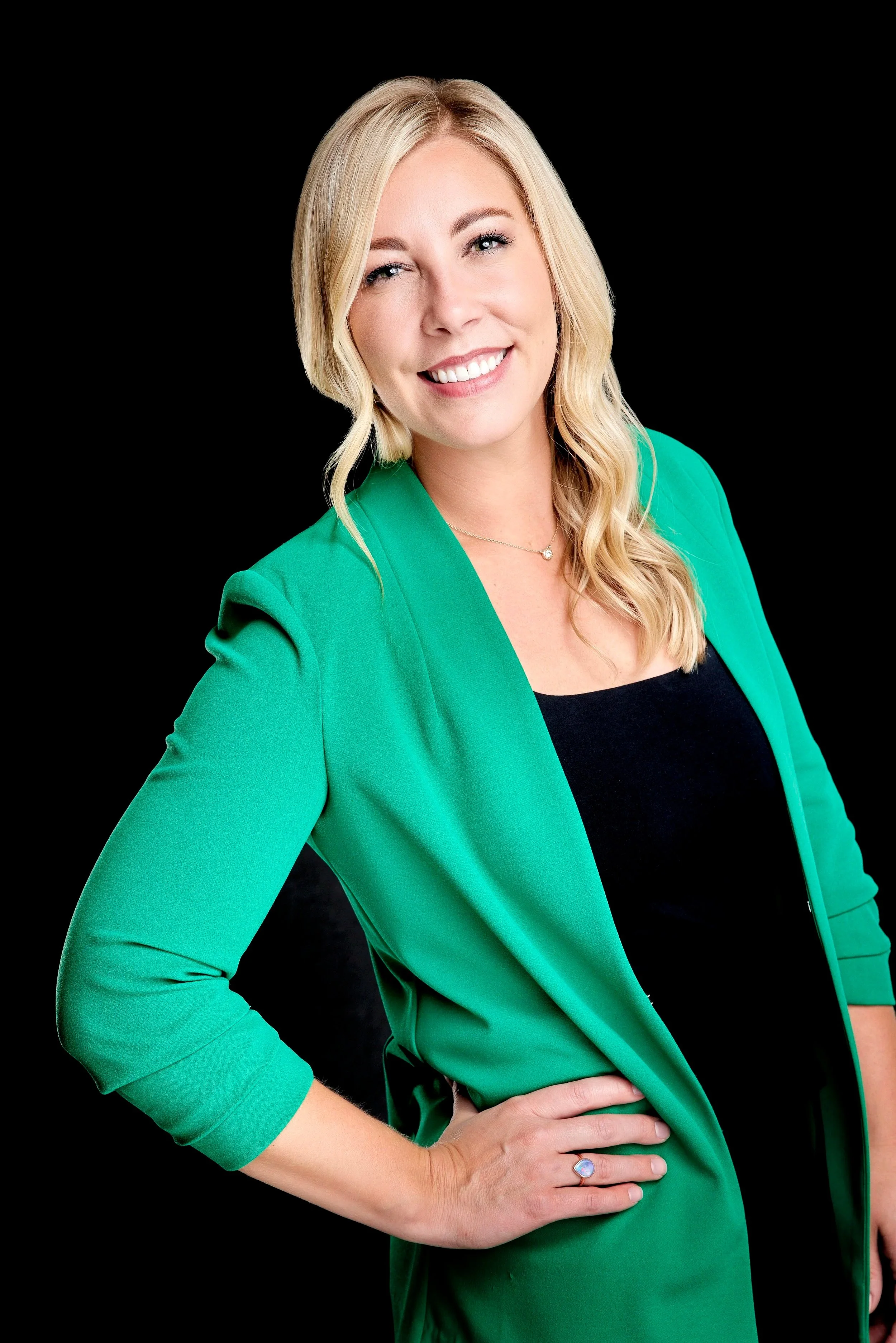 A woman with blonde hair wearing a green blazer over a black top, smiling and posing with her hand on her hip, against a black background.