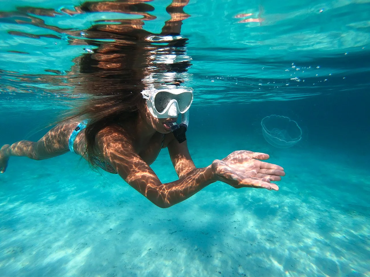 A woman snorkeling underwater, wearing a diving mask and snorkel, reaching out with one hand, with a jellyfish visible in the water nearby.