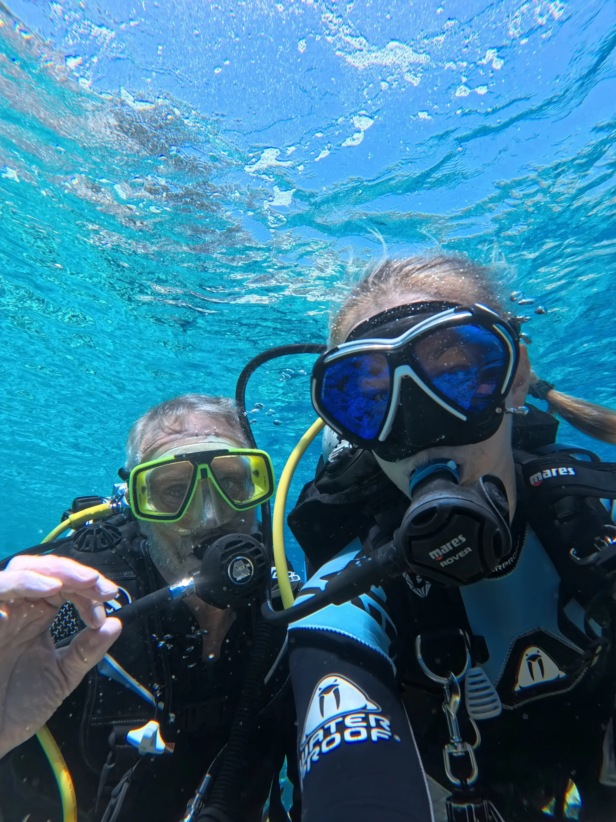 Two scuba divers underwater taking a selfie, wearing diving masks and gear, with blue water and bubbles around them.