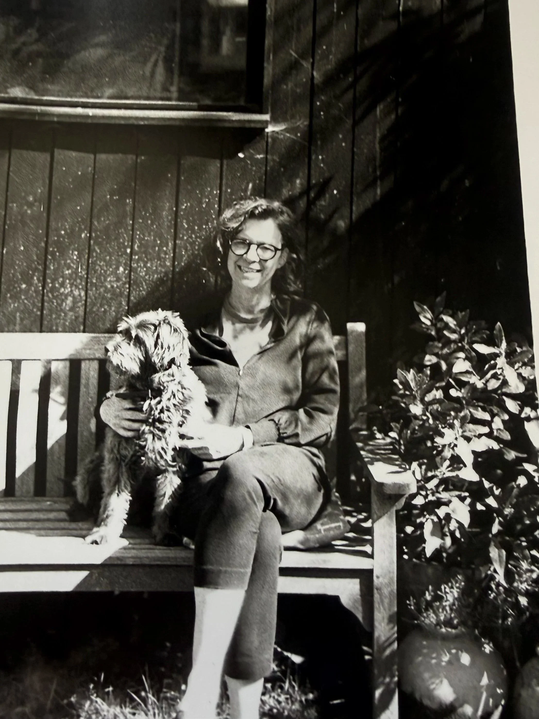 A woman with glasses and curly hair sitting on a park bench, holding a fluffy dog. There are plants and a tree with leaves in the background, and the photo is in black and white.