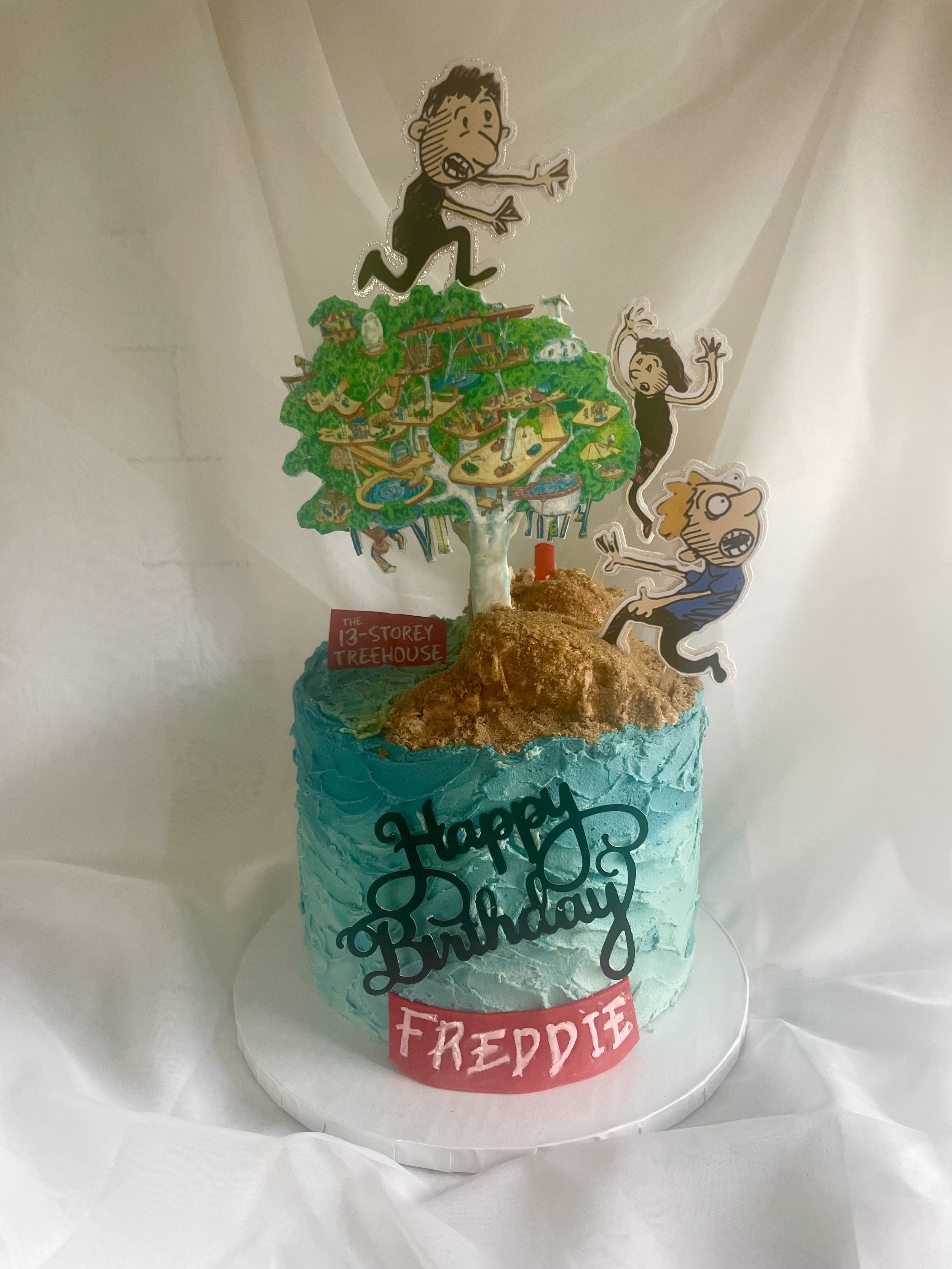 A birthday cake with blue frosting and a 'Happy Birthday Freddie' message, topped with a 'Peanuts' comic strip characters and a 'Treehouse' themed decoration.