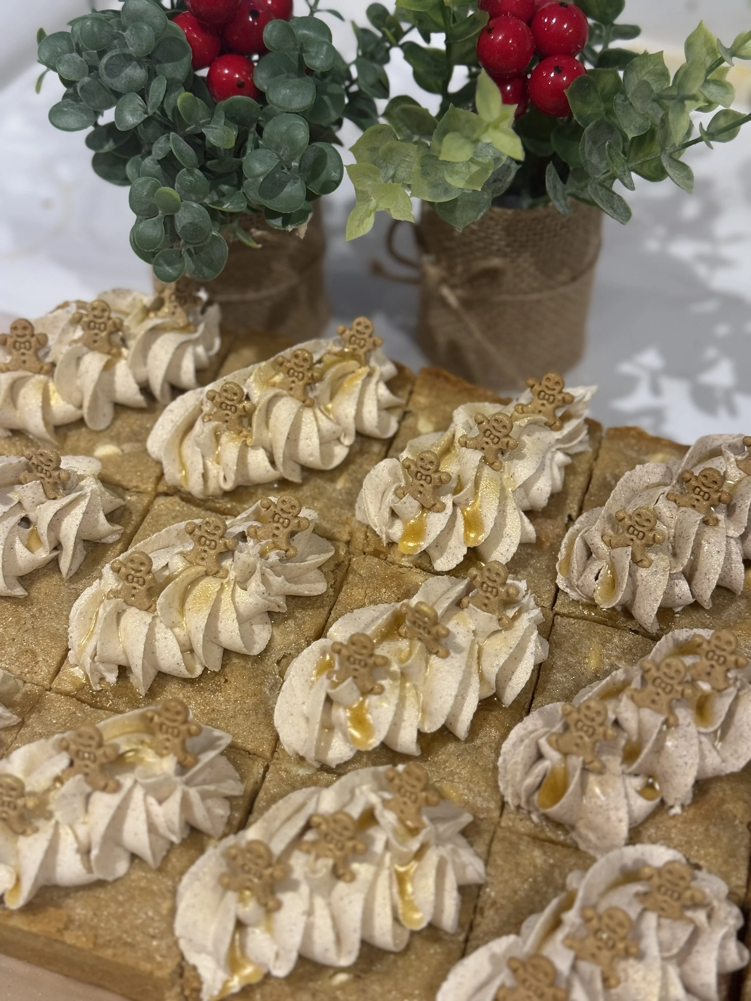 Assorted cakes topped with whipped cream, finger cookies, and gold syrup, arranged on a wooden tray, with potted holiday plants containing red berries, green leaves, and small brown paper-wrapped pots in the background.