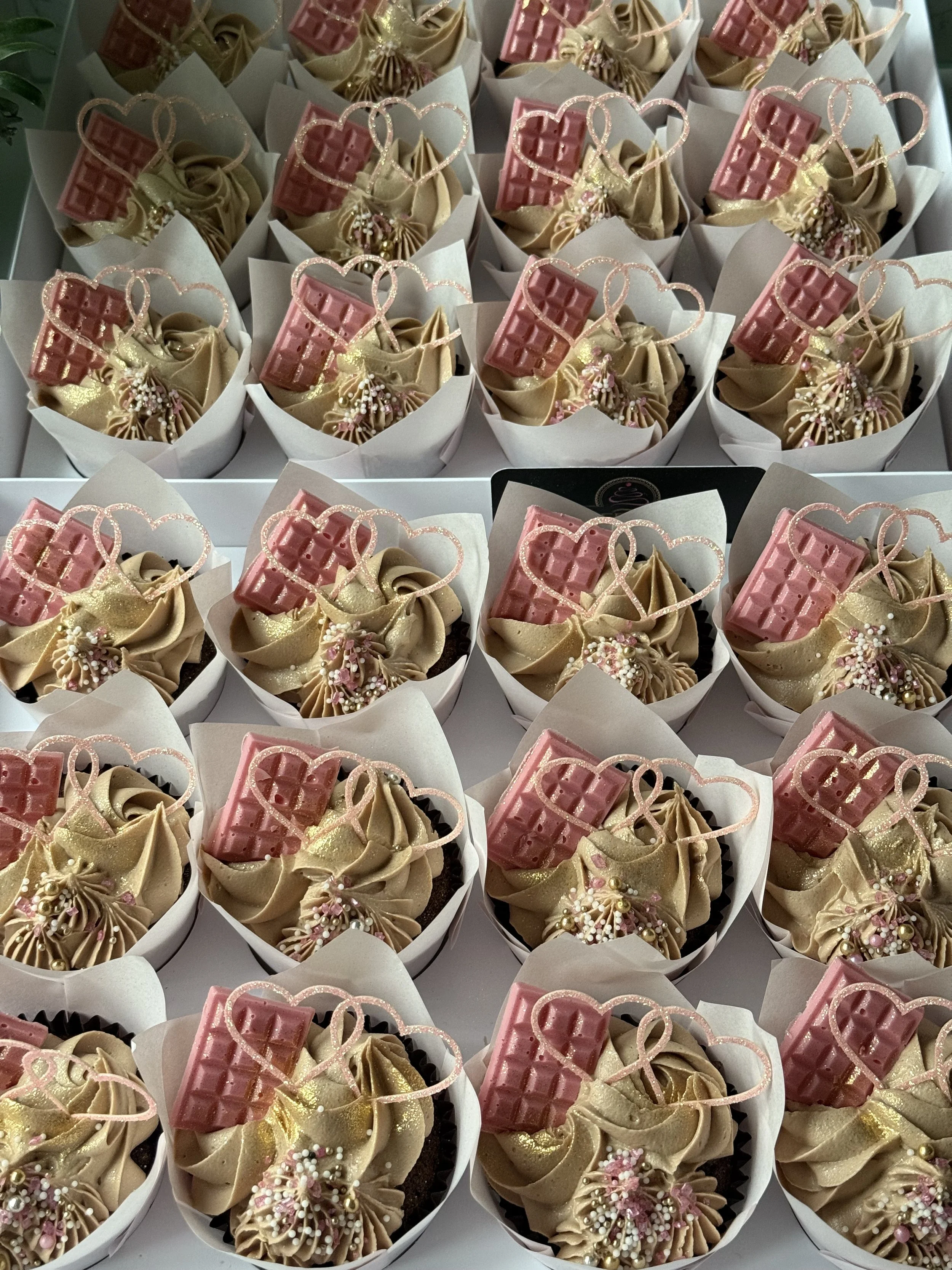 Multiple cupcakes decorated with pink chocolate bars, gold heart-shaped decorations, and sprinkles, arranged in white cupcake liners.