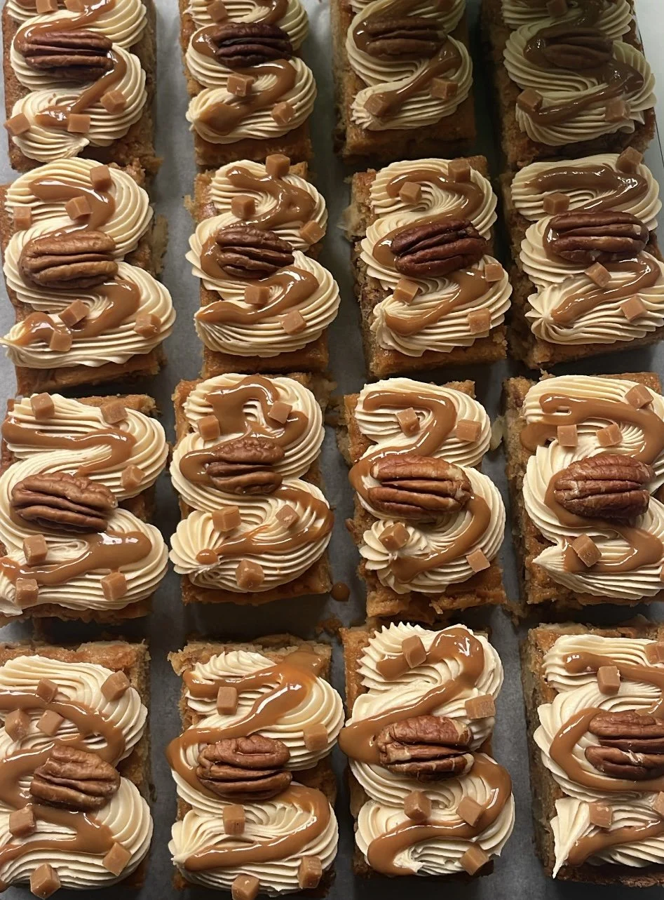 Row of square caramel and pecan desserts topped with whipped cream, caramel drizzle, pecans, and small caramel candies.