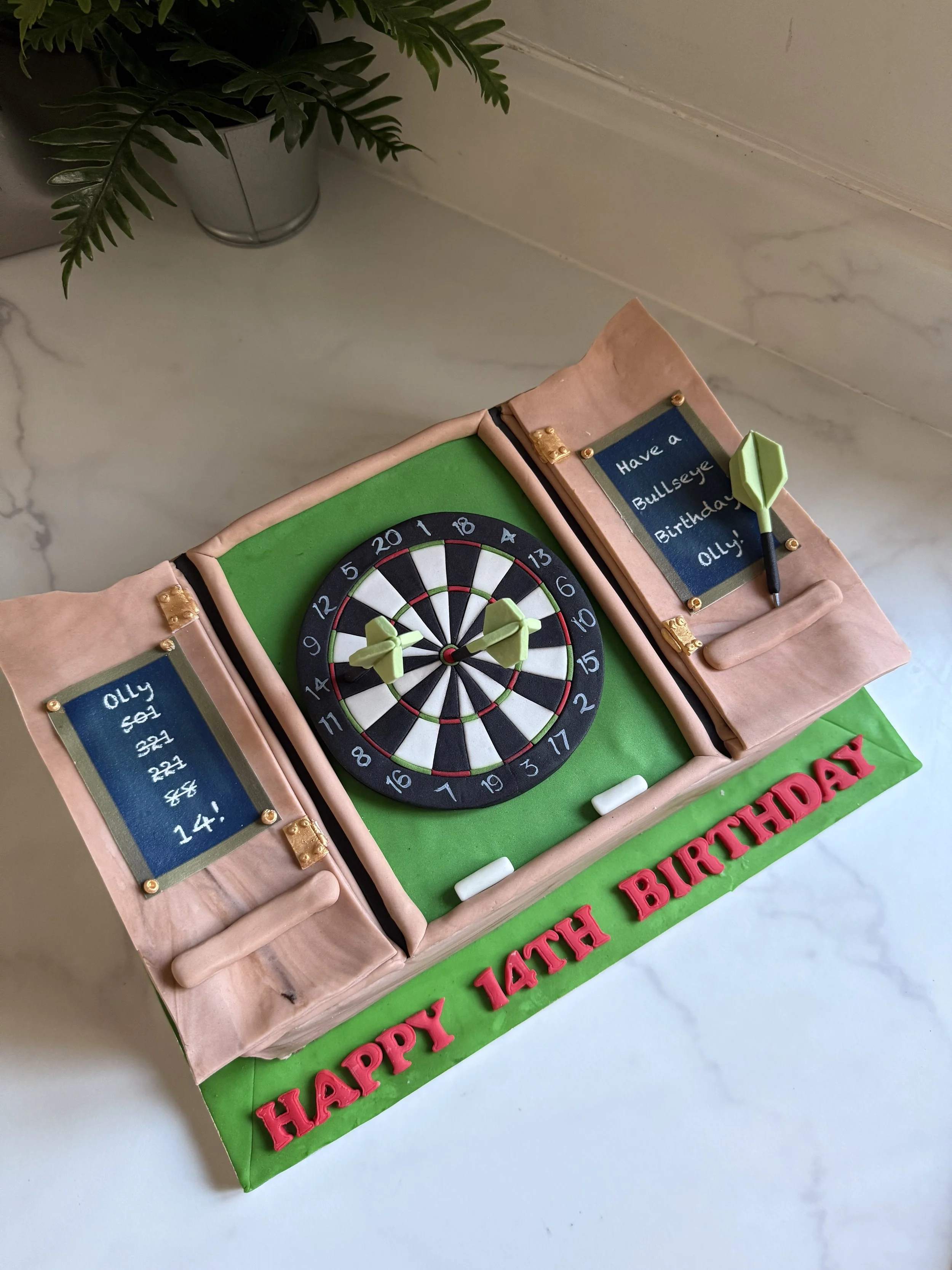 A birthday cake designed to look like a dartboard, with darts on it, and two scoreboards on either side with a birthday message. The cake has green and pink icing with red letters spelling 'HAPPY 14TH BIRTHDAY' on the base.