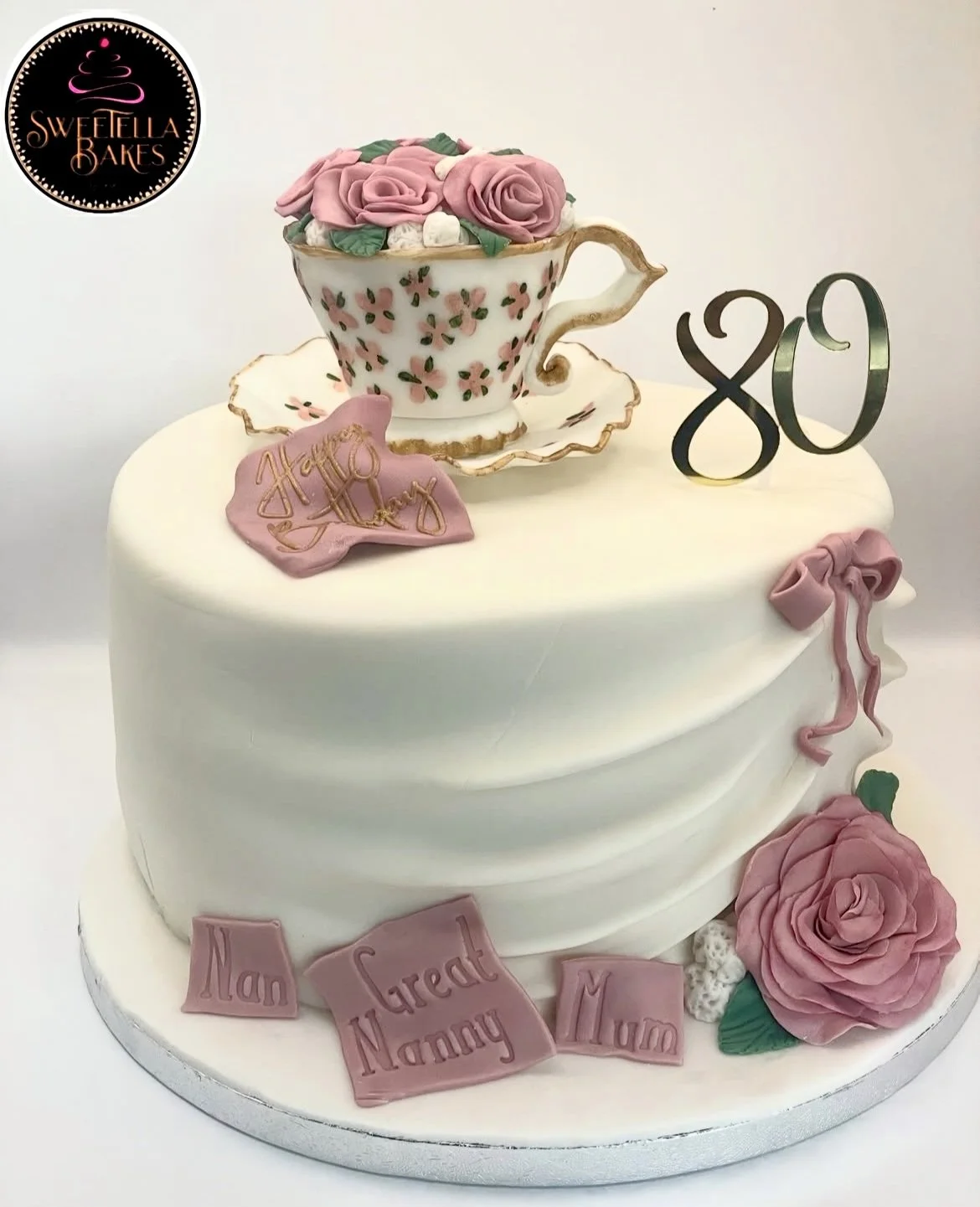 White cake with pink roses, a teacup with roses on top, and a 'happy 80th' decoration. The cake has pink and white fondant accents, including roses, a bow, and pink plaques with 'Nan', 'Great', 'Nanny', and 'Mum' written on them. The logo in the top 