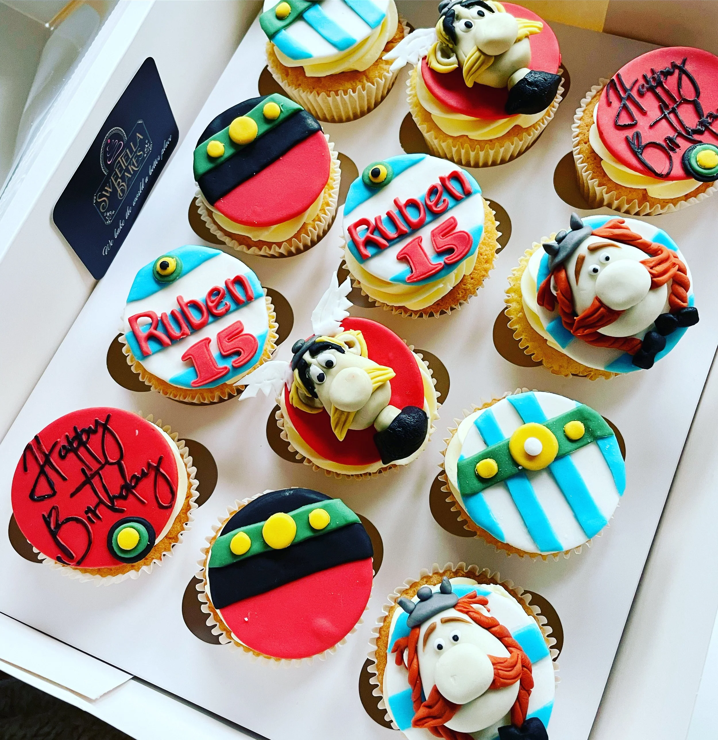A display of colorful birthday cupcakes with various decorations, including characters and text, arranged on a white surface.