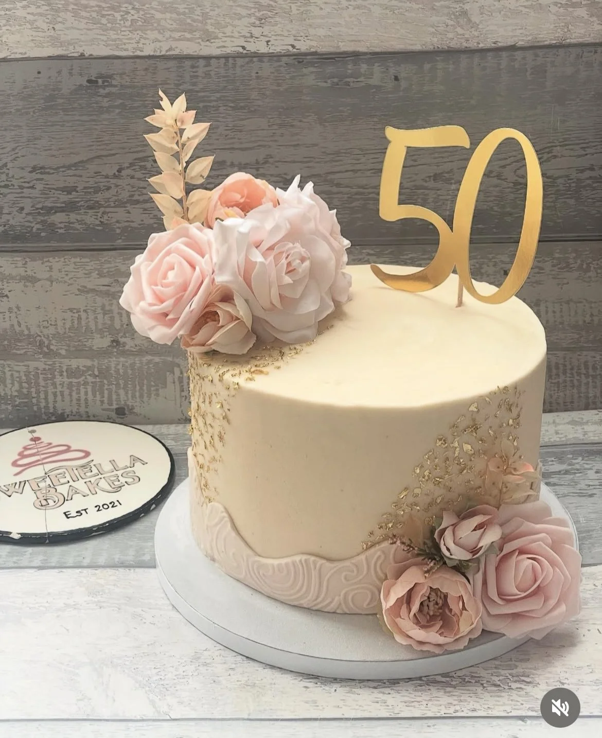 A white birthday cake decorated with pink and cream roses, gold accents, and a gold topper with the number 50, placed on a white cake board.