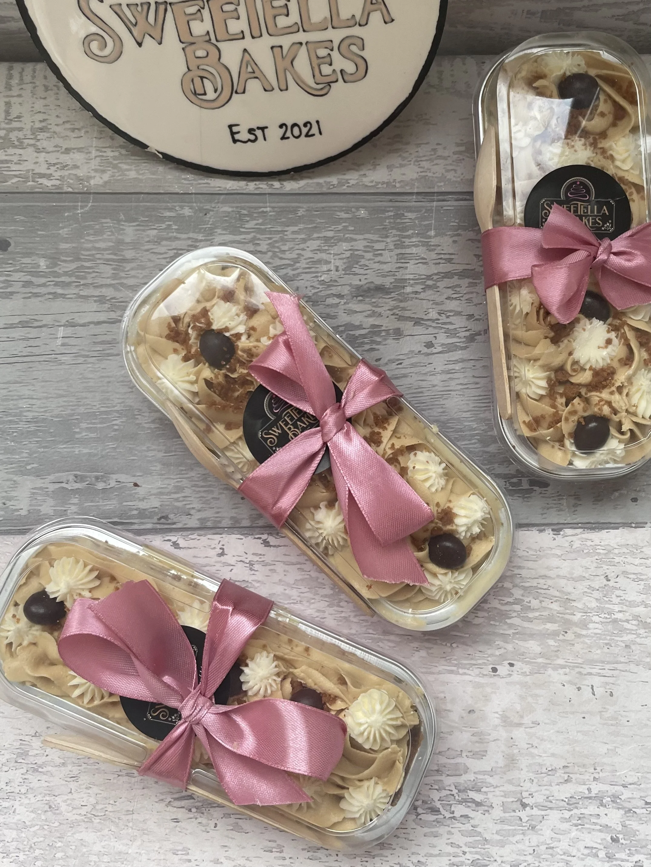 Three clear plastic containers of decorated cake with pink ribbons on a gray wooden surface, with a round sign that reads 'SWEETIELLA BAKES EST 2021' in the background.