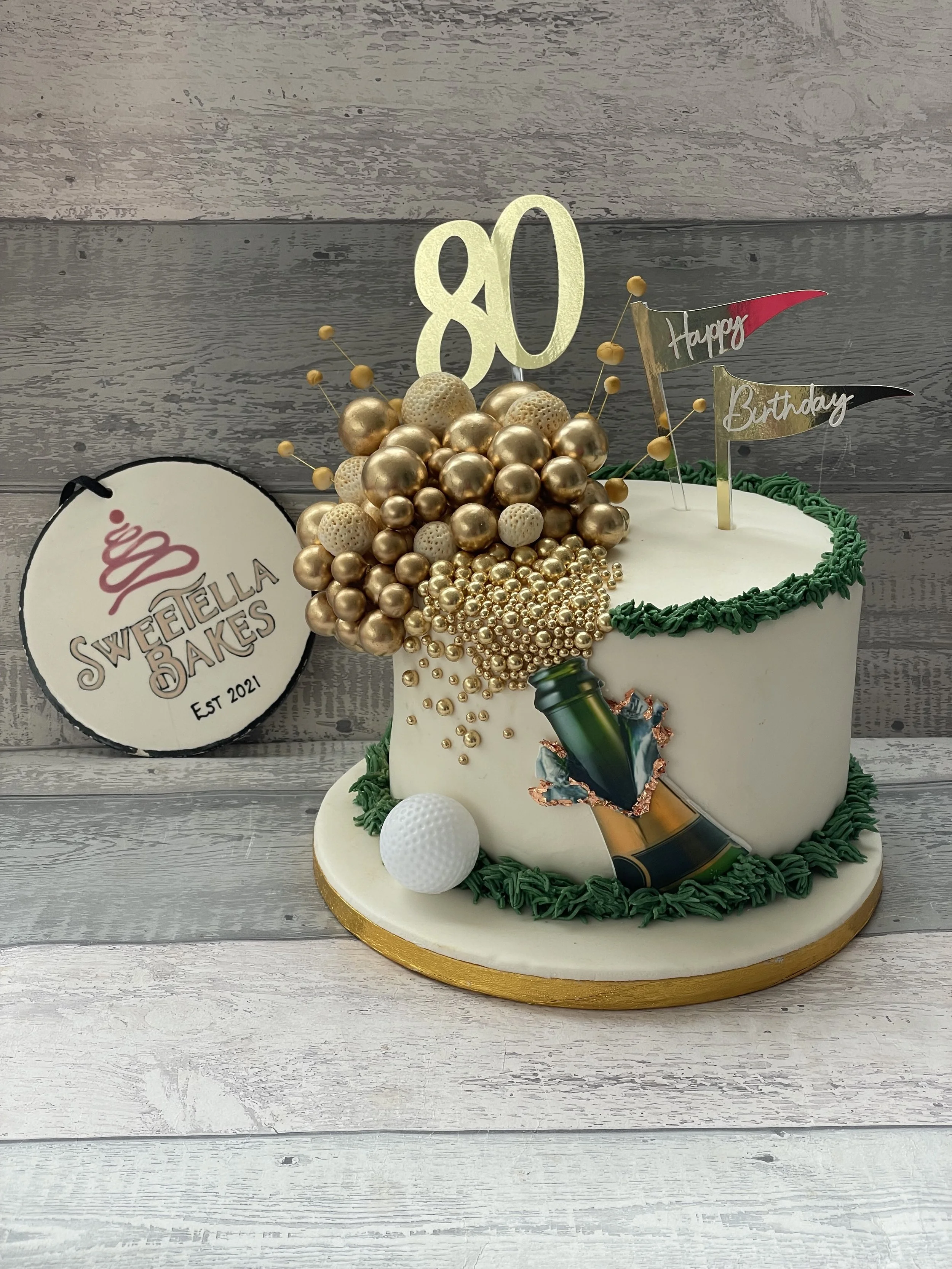 A birthday cake with a white base decorated with green icing, gold and white spherical decorations resembling a champagne bottle and bubbles, and a gold '80' topper. There are two small flags reading 'Happy' and 'Birthday.' A golf ball is placed on t