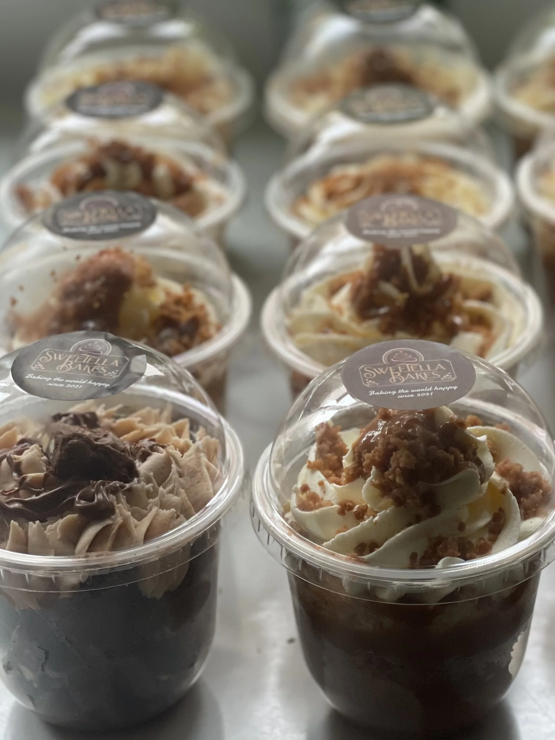Several clear plastic cups filled with assorted layered desserts topped with whipped cream and crumbled toppings, labeled with a sticker from 'Sweetella Bakes'.