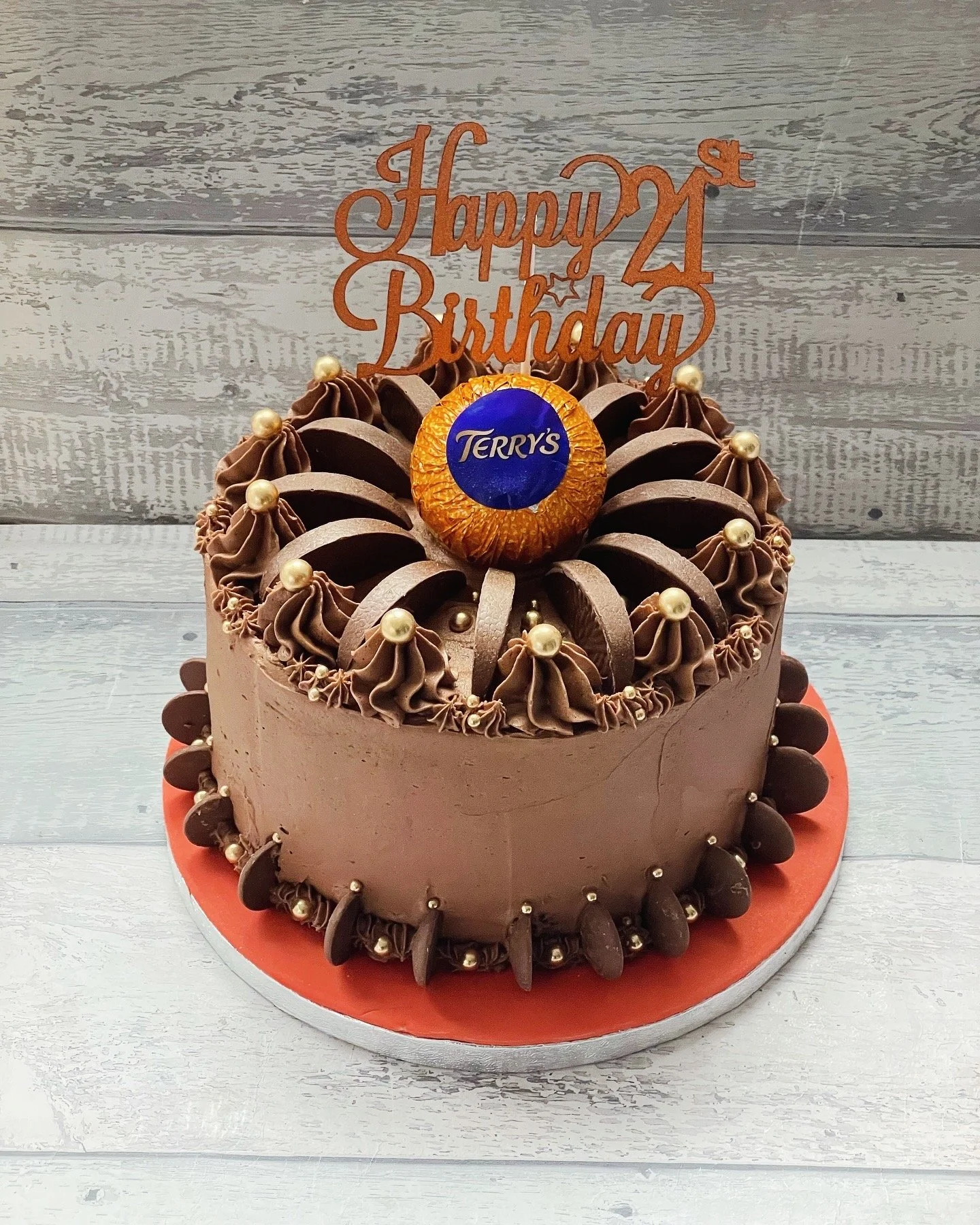 Chocolate birthday cake with decorative chocolate icing, gold pearls, and a large chocolate candy in the center. The cake has a red 'Happy 24th Birthday' topper and is on a silver cake board.