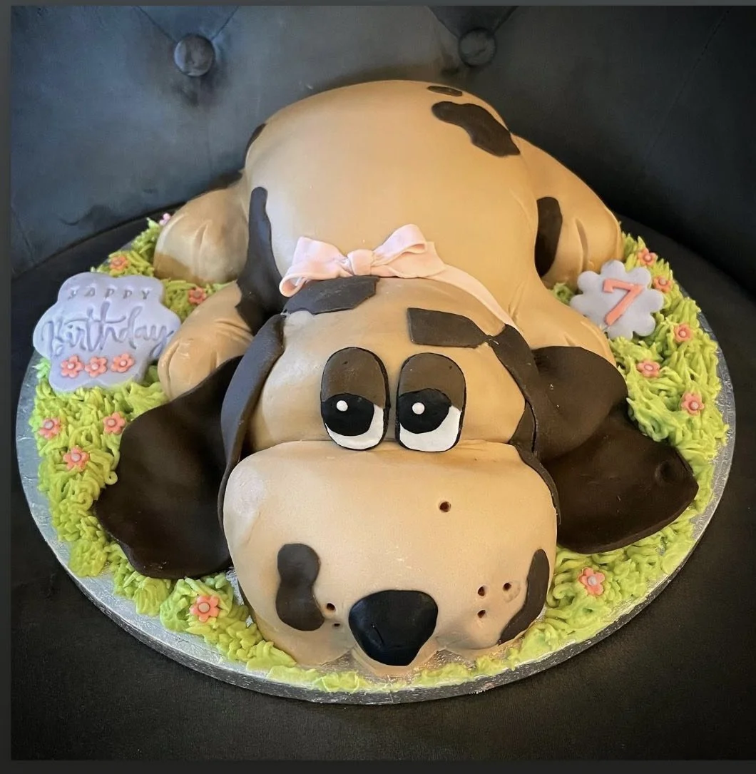 Cake decorated as a cute dog lying on its back with a pink bow around its neck, placed on a bed of green frosting with pink flowers, celebrating a 7th birthday.