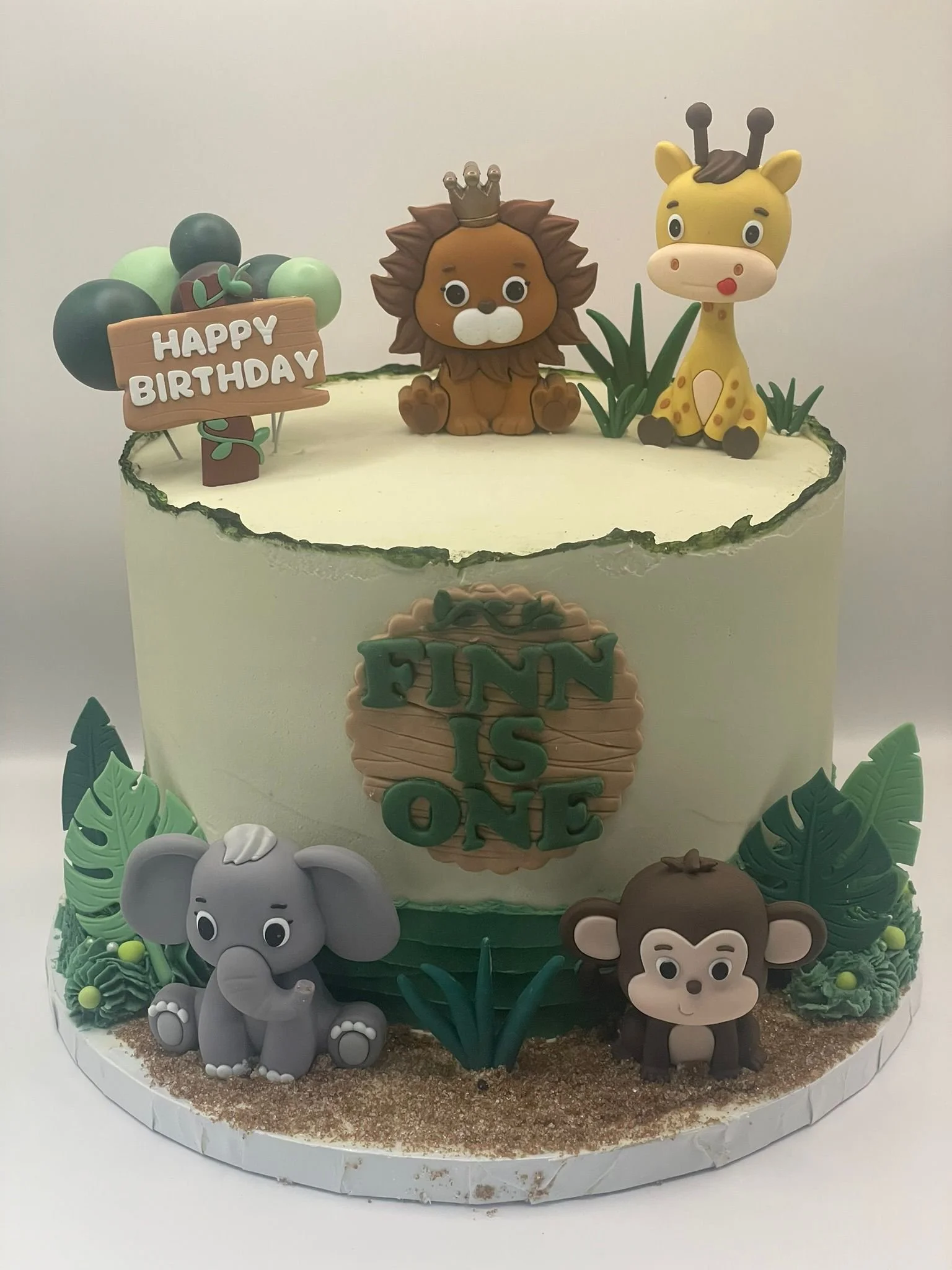Animal-themed birthday cake with figurines of a lion, giraffe, elephant, and monkey, decorated with jungle leaves and a sign that reads 'Happy Birthday' and 'Finn is One'.