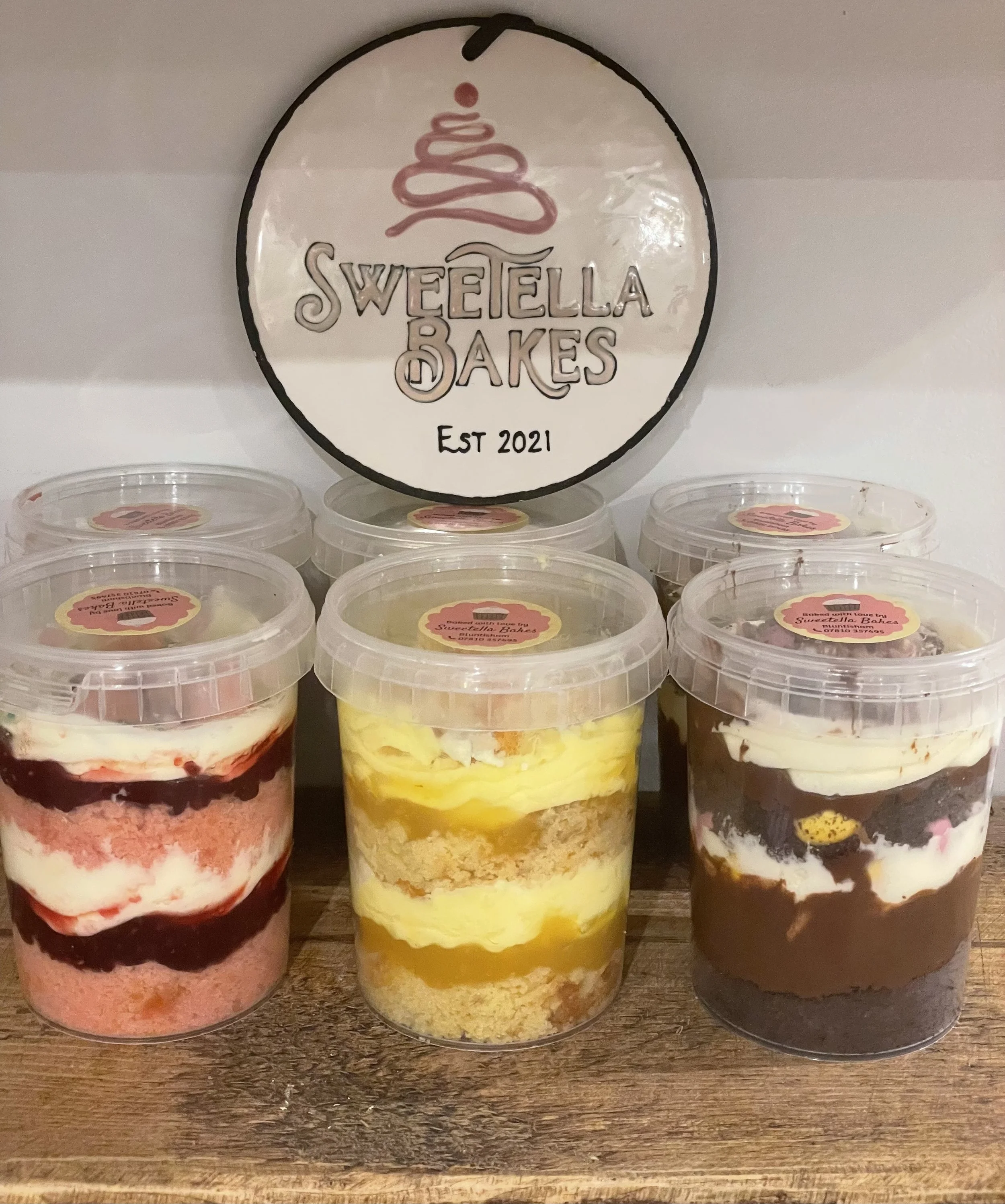 Assorted jars of layered cakes from Sweettella Cakes on a wooden surface with a circular sign in the background that says 'Sweettella Cakes Est 2021'.