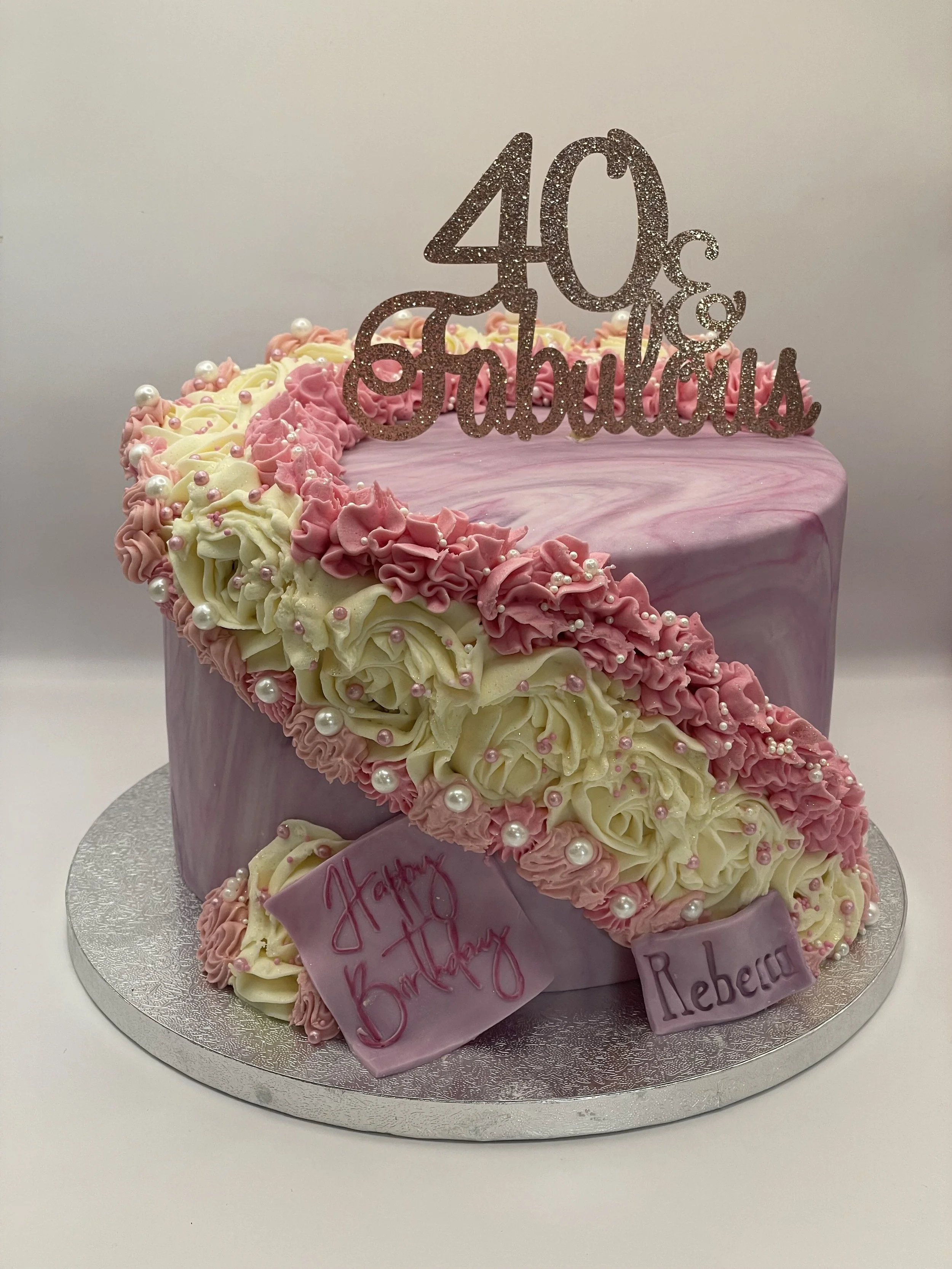 Pink and cream decorated birthday cake with a '40 and Fabulous' glitter topper, pink and white icing roses, and edible pearls. There are two pink plaques with 'Happy Birthday' and 'Rebeca' written on them. The cake is on a silver cake board.