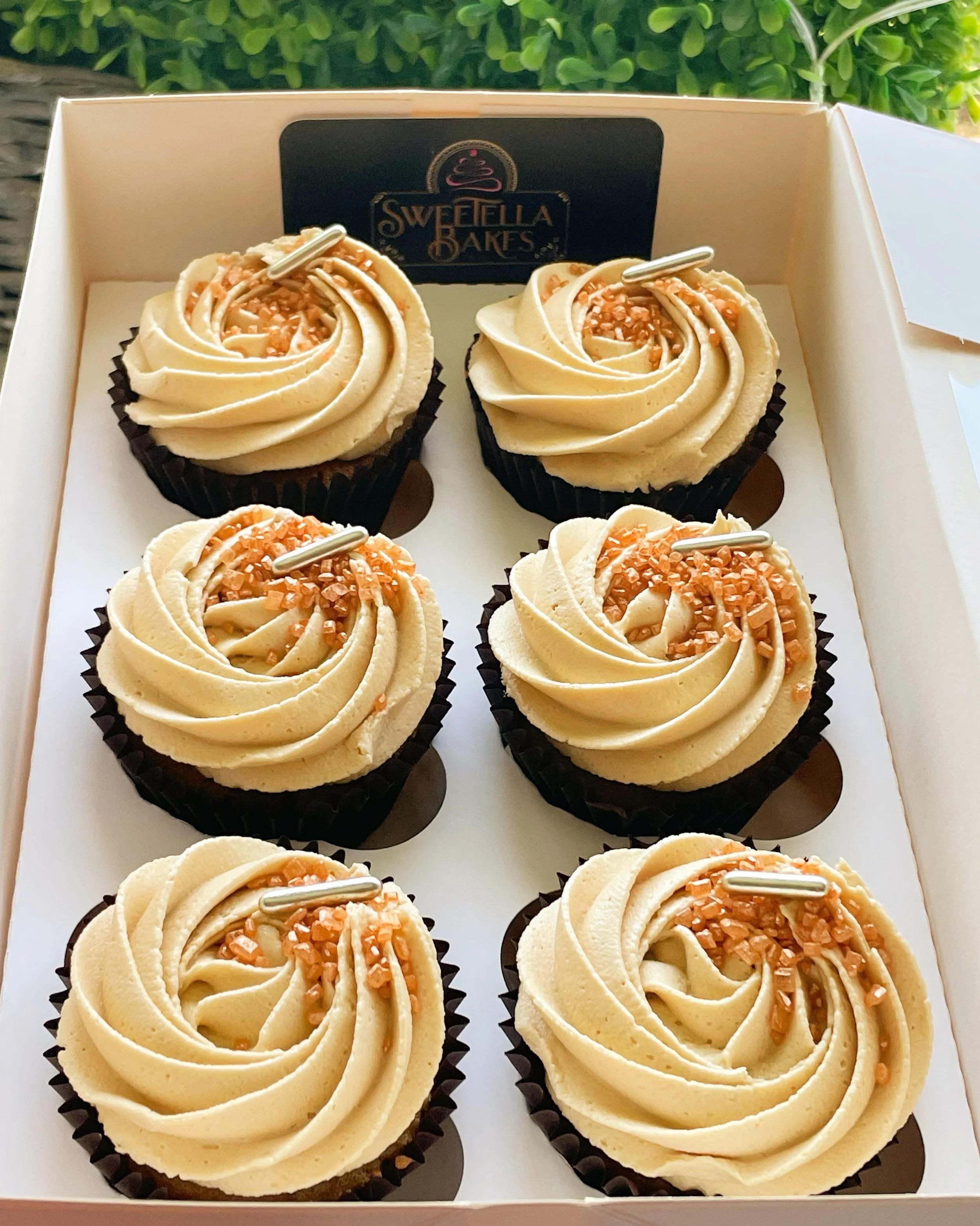 Six cupcakes with cream-colored frosting topped with chopped nuts and a small silver decoration, in a white box with a black label reading 'Sweetella Bakes'.