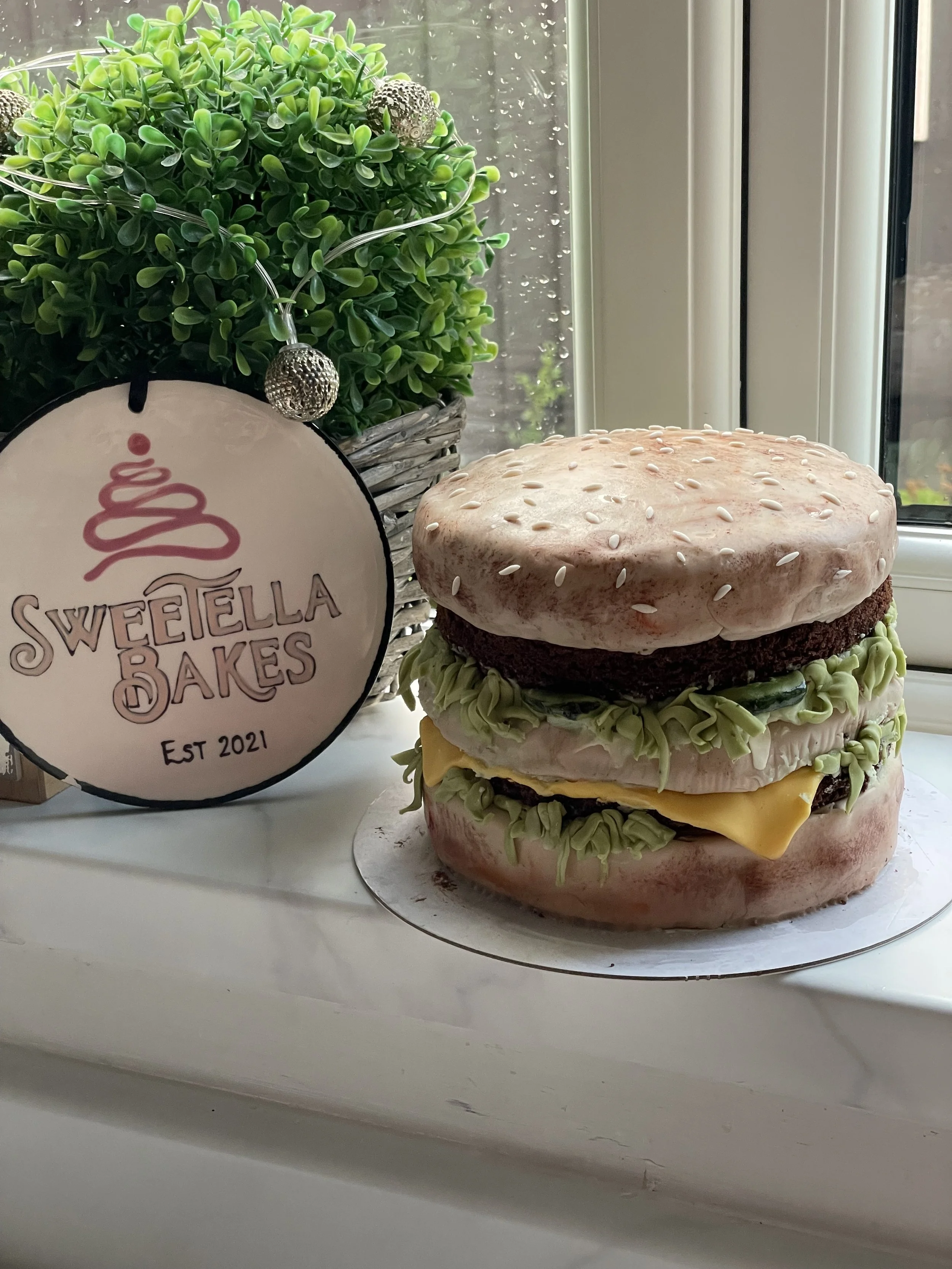 Cake shaped to resemble a hamburger with bun, lettuce, cheese, and burger patty, placed on a white surface near a potted plant with a Sweeella Bakes sign hanging from it, inside a room near a window.
