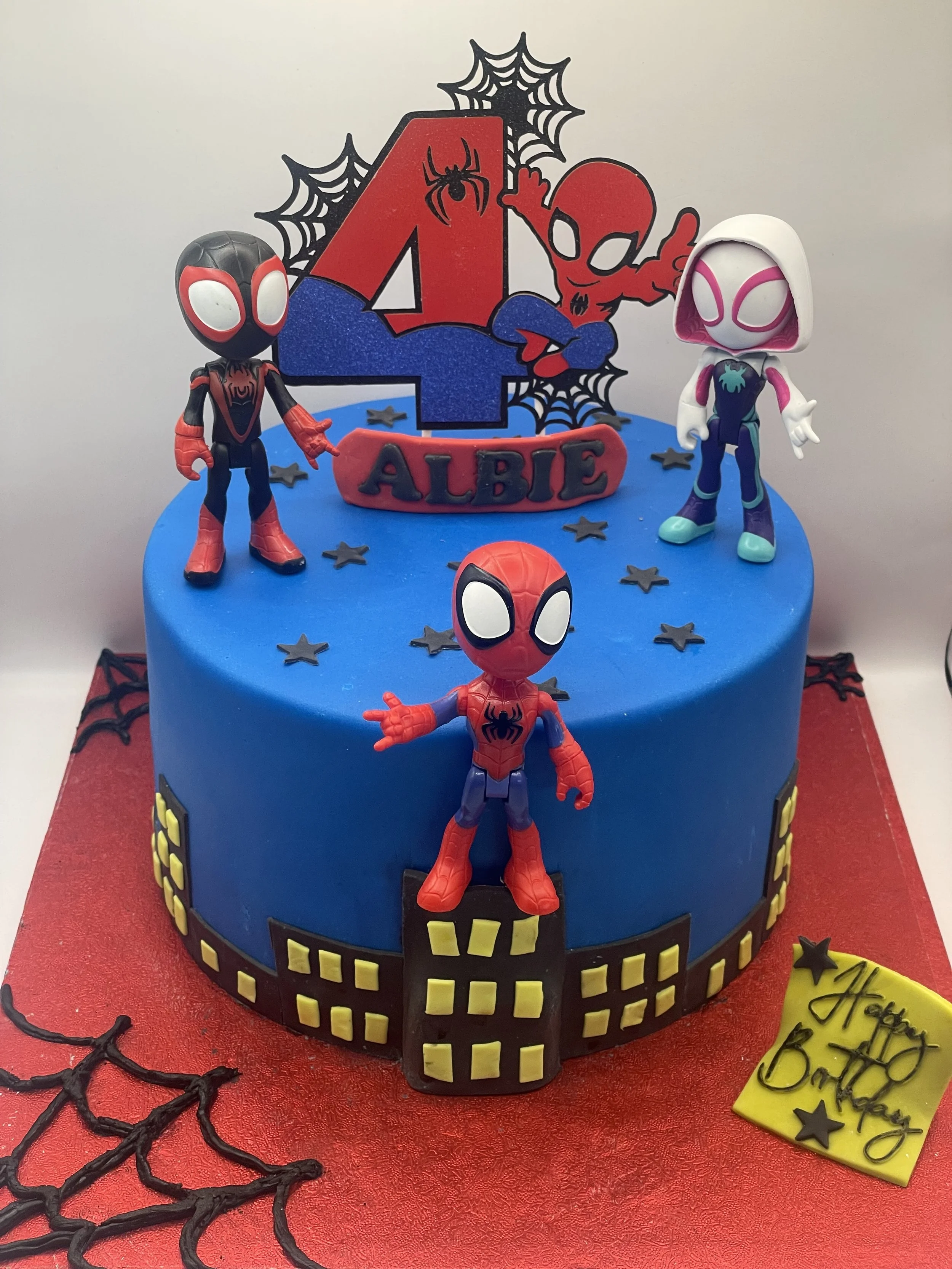 Spider-Man themed birthday cake with three Spider-Man figurines on top, blue cake with yellow windows, spider webs decoration, and a sign that reads 'Happy Birthday' on a red cake board.