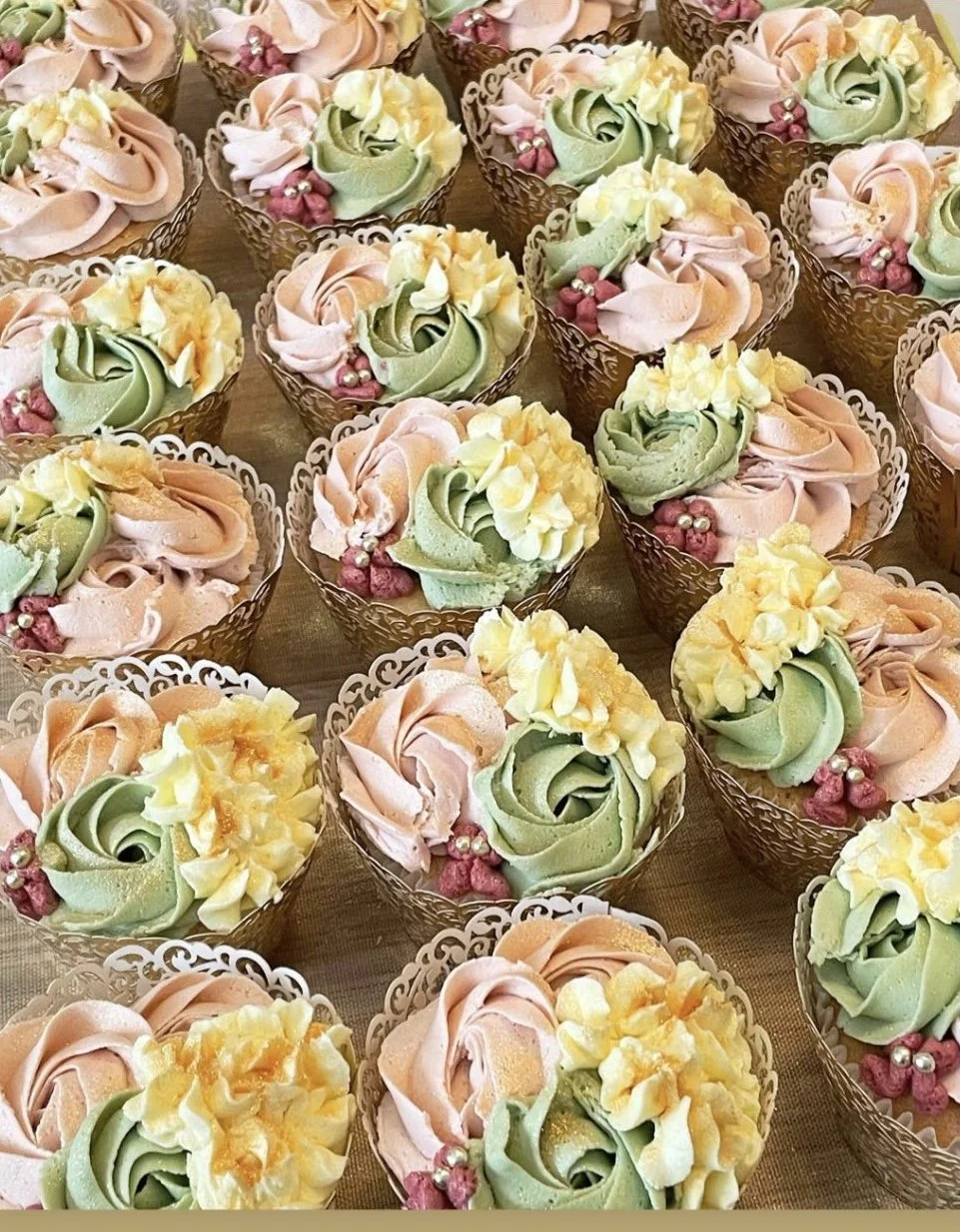 Assorted decorated cupcakes with pastel-colored frosting in pink, green, and yellow, topped with small decorative pearls and edible flowers, arranged in metallic wrappers.
