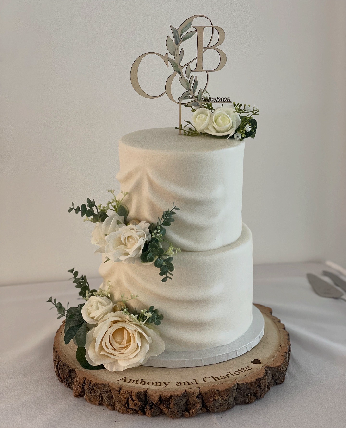 Two-tier white wedding cake decorated with white roses and green foliage, placed on a wooden slab with the names "Anthony and Charlotte" engraved, topped with a cake topper featuring initials and a date.