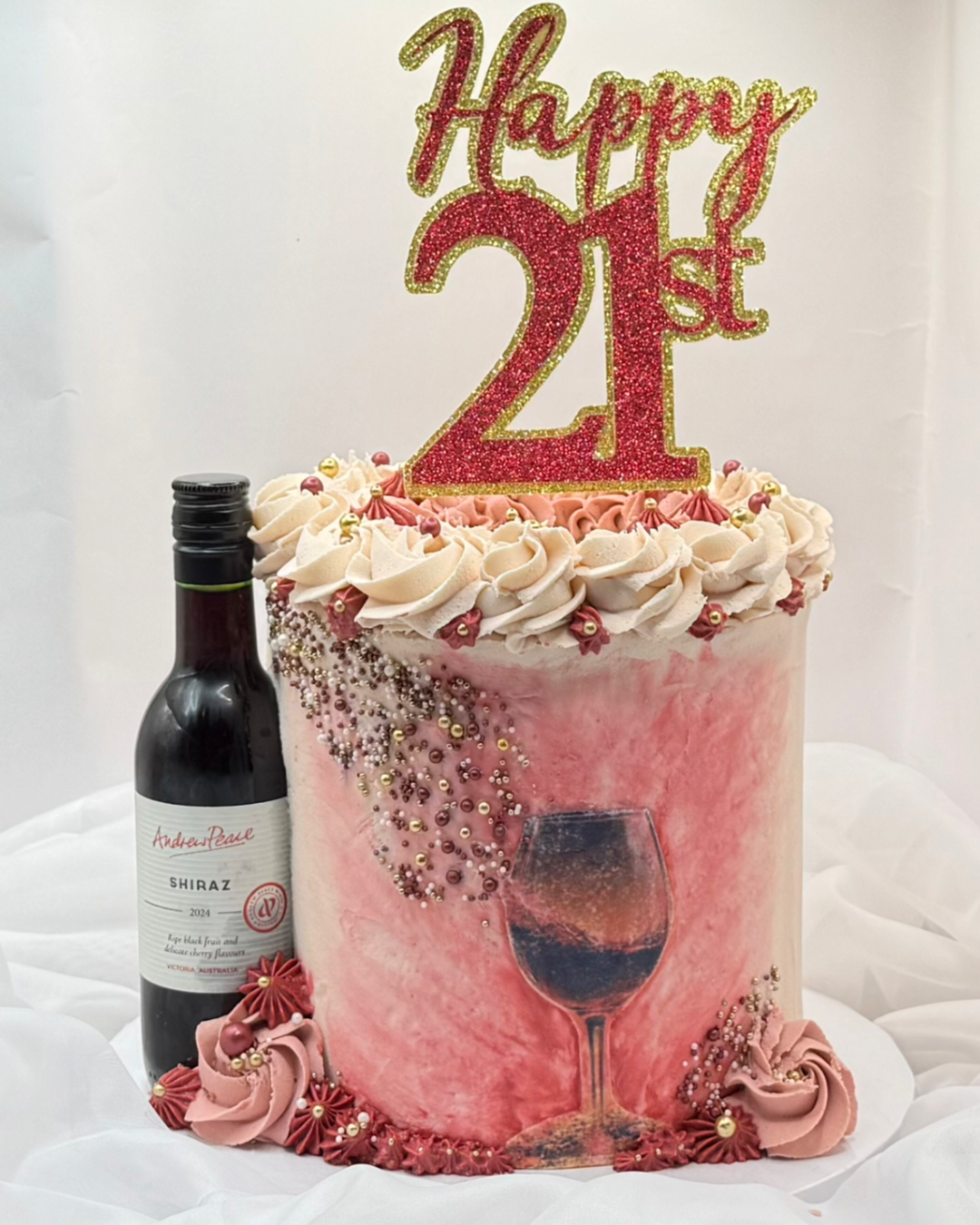 A festive birthday cake with pink and white marbled icing, decorated with pink and beige icing rosettes, pearl sprinkles, and gold beads. A glittery red and gold 'Happy 21st' topper is placed on top. The cake features a painted image of a wine glass 