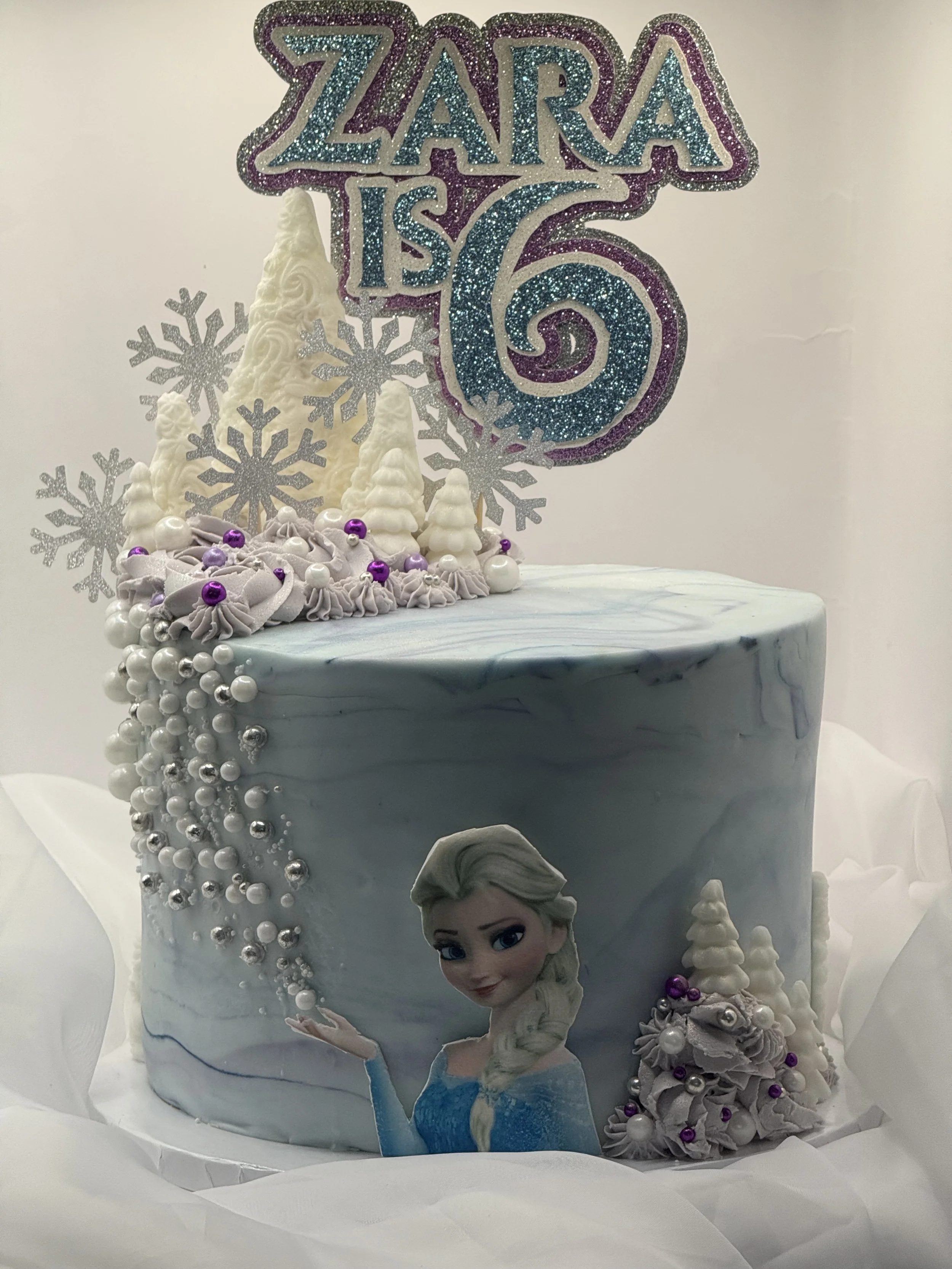 A Frozen-themed birthday cake for a girl named Zara celebrating her sixth birthday. The cake features an image of Elsa from Frozen, silver and purple decorations, fondant snowflakes, and a glittery sign that reads "Zara is 6."