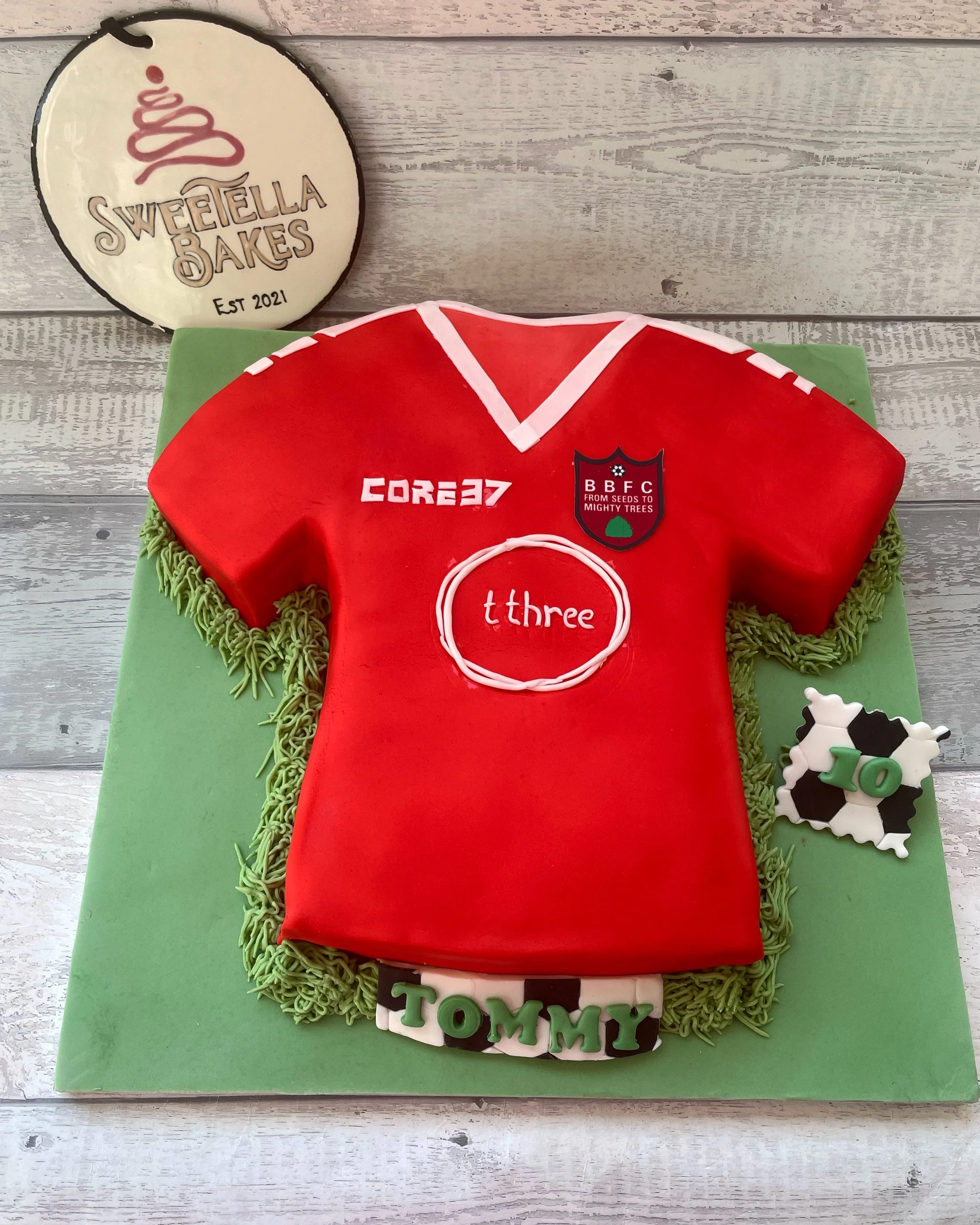 A cake shaped like a red sports jersey with white icing accents, green piping, and a custom name "TOMMY" at the bottom. The cake features sports logos and the number "3" in the center, and decorations including a soccer ball with the number "10" and 