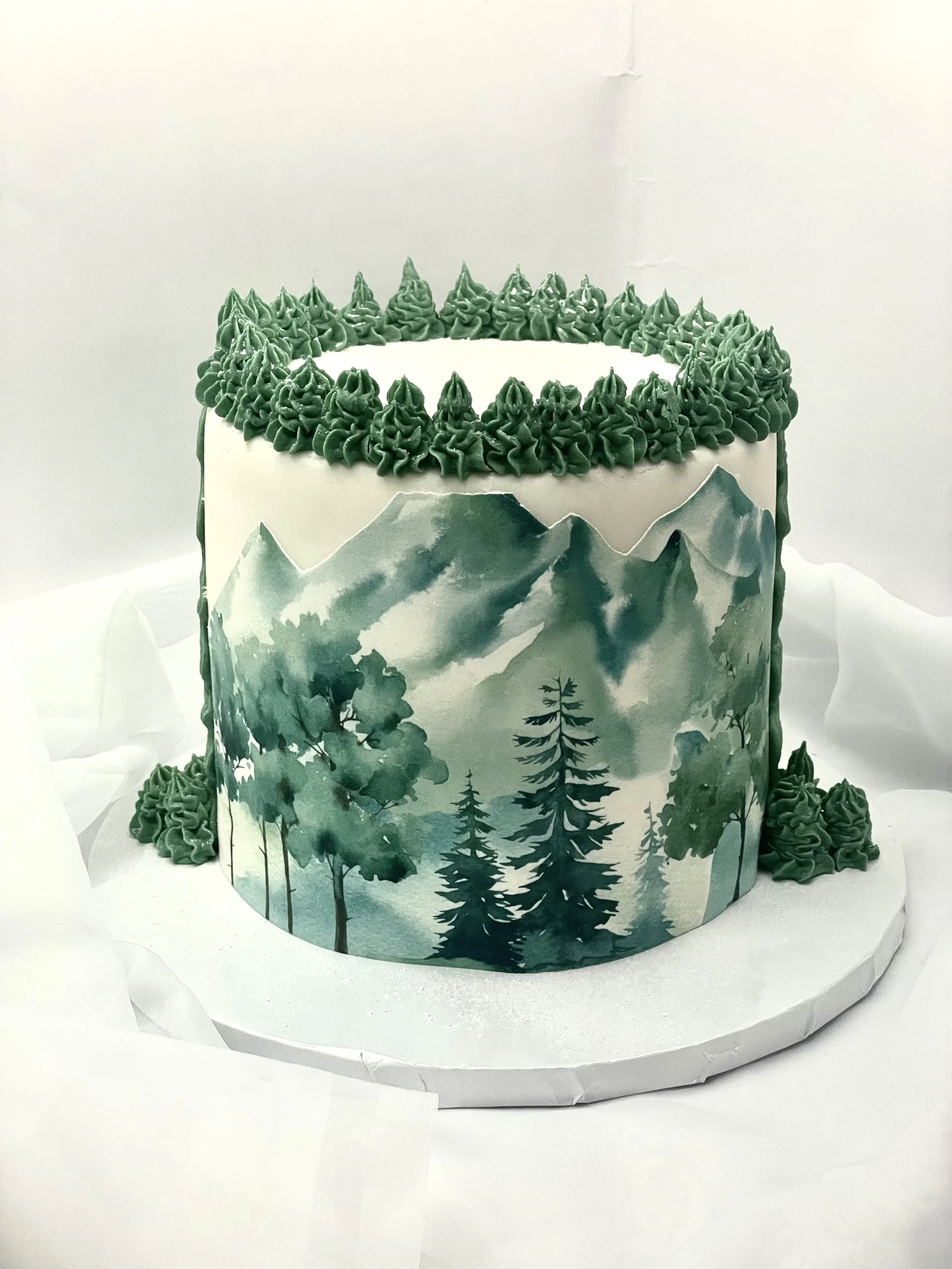 A cake with mountain and forest landscape design, decorated with green piped frosting on top and sides, placed on a white round base.