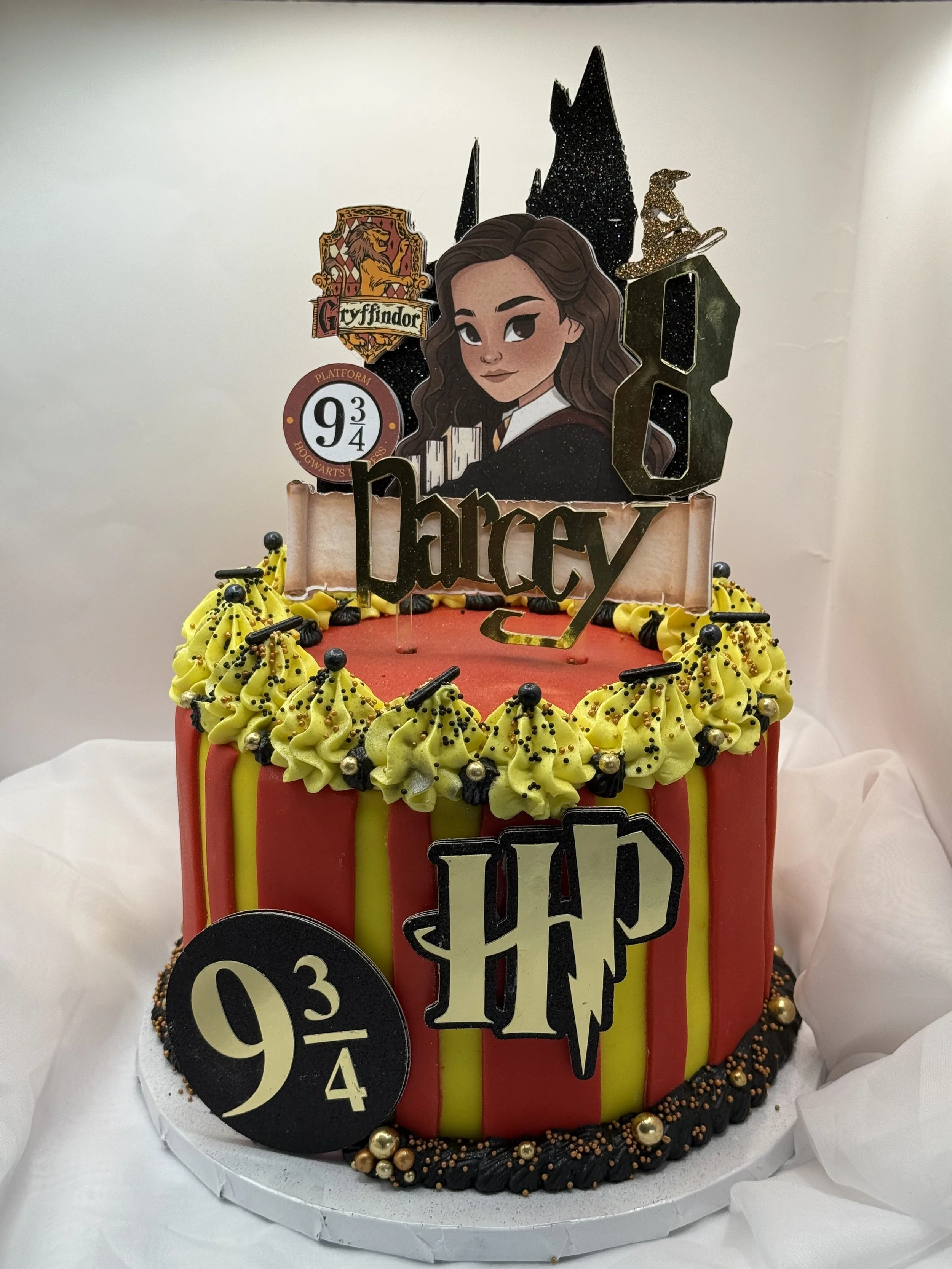 Harry Potter-themed birthday cake with red and yellow striped icing, black and gold decorations, and Harry Potter and Harry Potter-themed toppers including a caricature of Hermione Granger, the Hogwarts crest, and the Number 8.