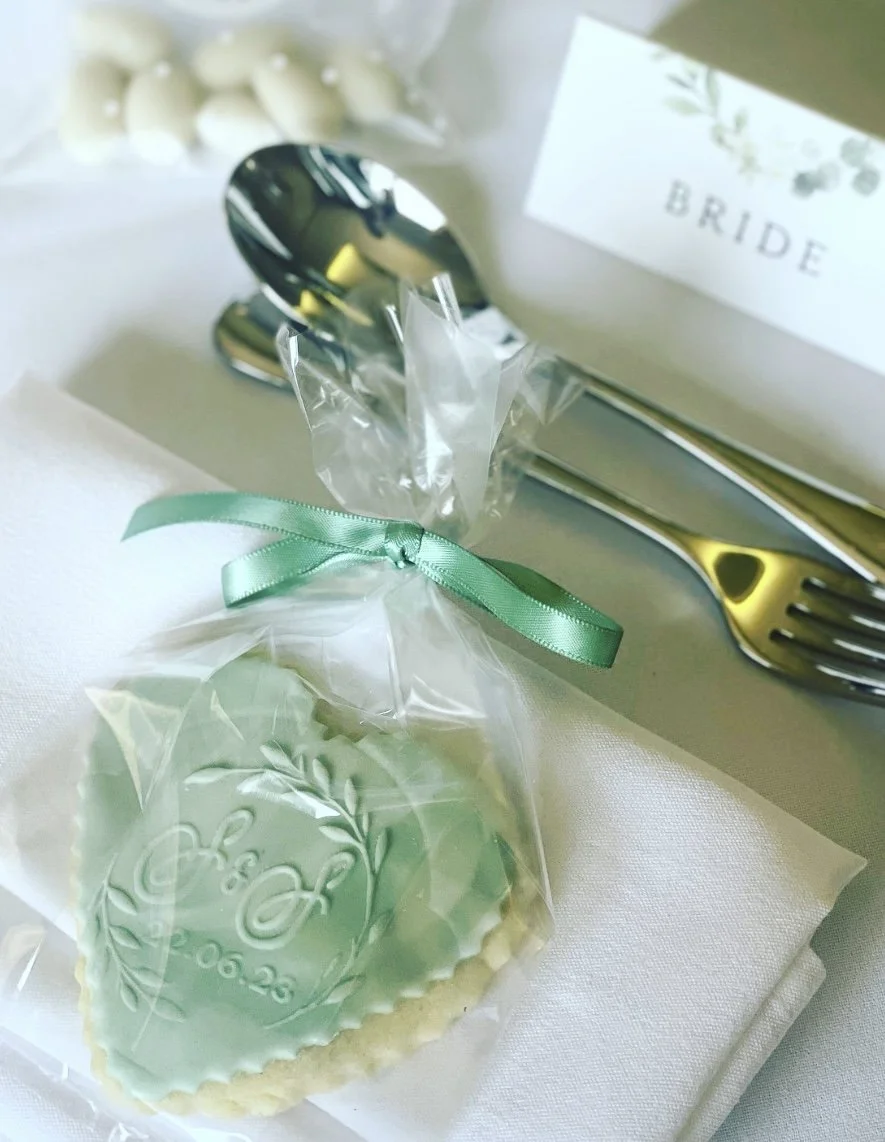 A wedding favor of a decorated cookie with an inscription 'Cherish' in a plastic bag tied with a green ribbon, a silver spoon, a 'Bride' card, and some white decorative beads in the background.