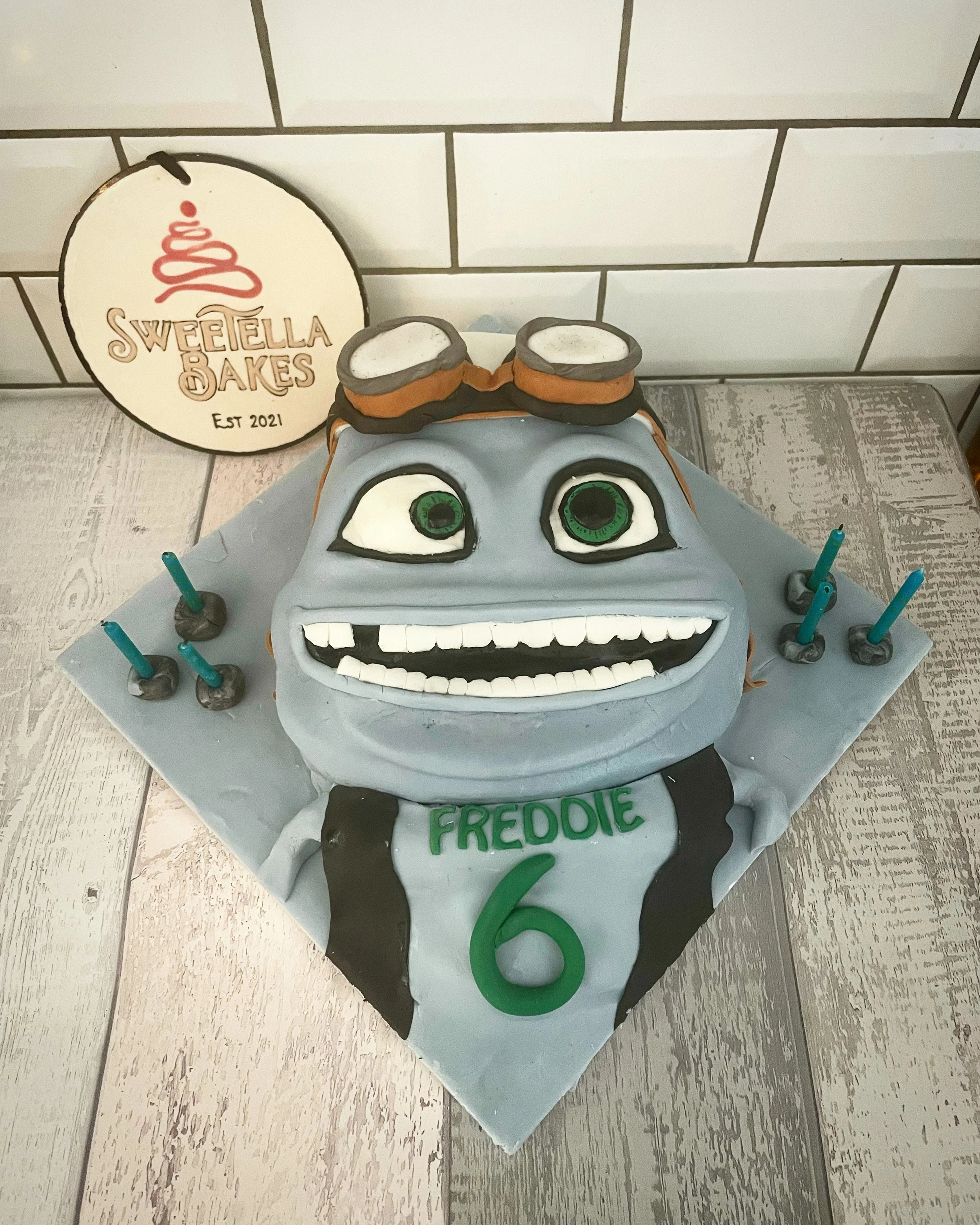 A cake designed to look like a cartoon character with big green eyes, a wide smiling mouth with white teeth, and gray skin. The cake has a black and gray baseball jersey with the name 'FREODIE' and the number '6' on it, and it is decorated with small