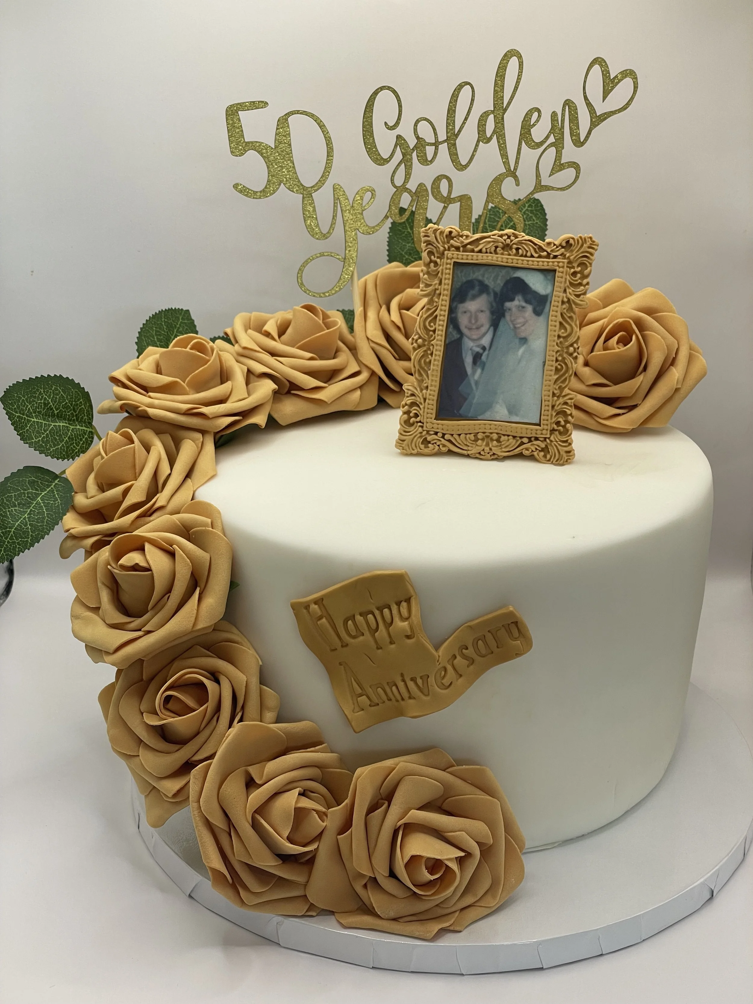 A white anniversary cake decorated with peach-colored roses, green leaves, a vintage photo frame with a picture of a couple, and a gold glittery sign that says '50 Golden Years' along with a gold banner that says 'Happy Anniversary'.