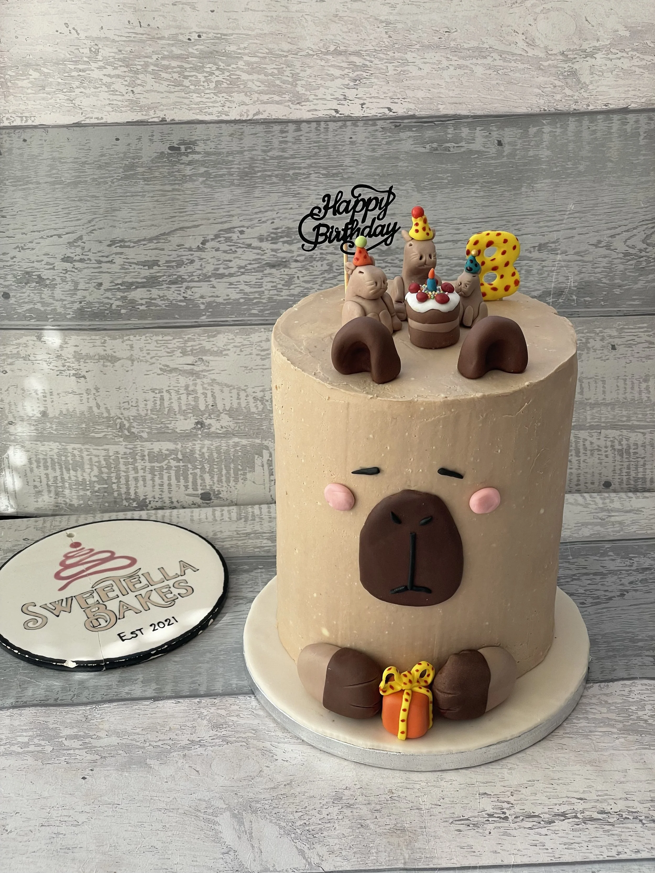 A birthday cake in the shape of a bear with a cute face, paws, and ears, decorated with fondant and animal-themed toppers including small bears with party hats, a number 3 candle, and a 'Happy Birthday' sign. The cake is placed on a white cake board 