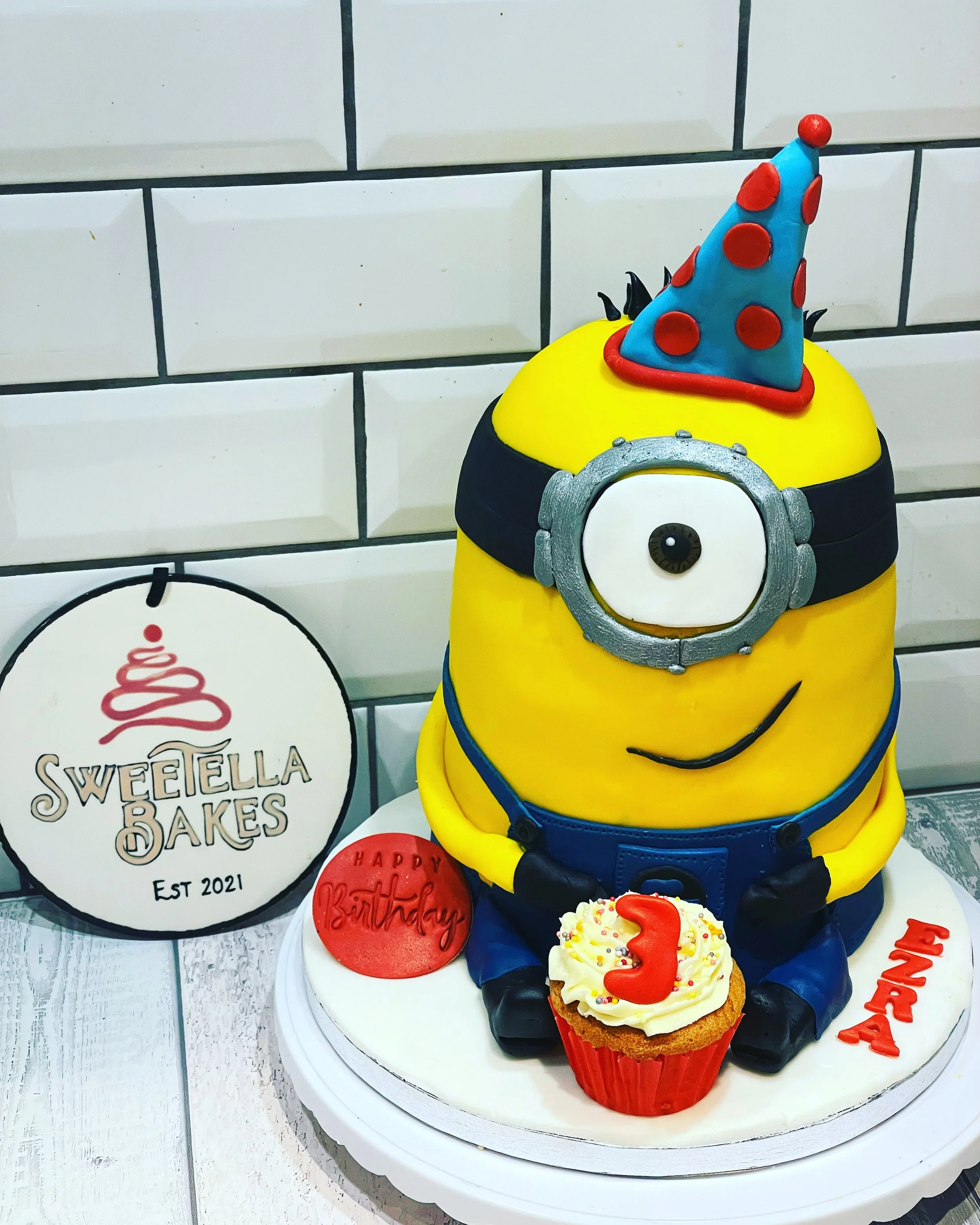 A Minion-themed birthday cake with one eye, a blue party hat with red polka dots, black gloves, and blue overalls, sitting on a white cake board with a cupcake topped with a red number '3' and a circular red birthday message plaque. There is a sign t