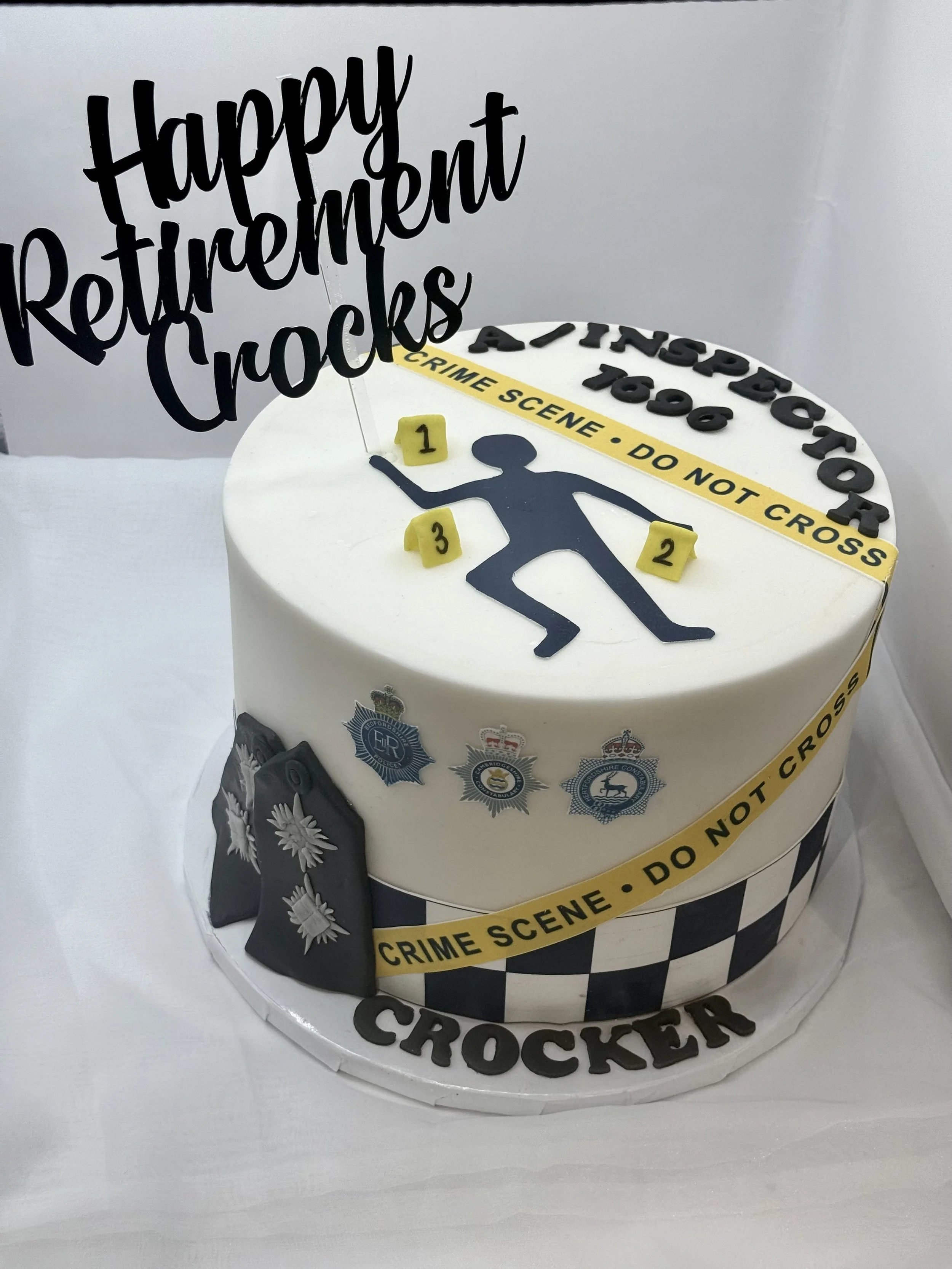 A birthday cake decorated with police-themed crime scene tape, badges, and police hats. The cake features a silhouette of a person with numbered evidence markers and a sign that says "Happy Retirement Crocket."