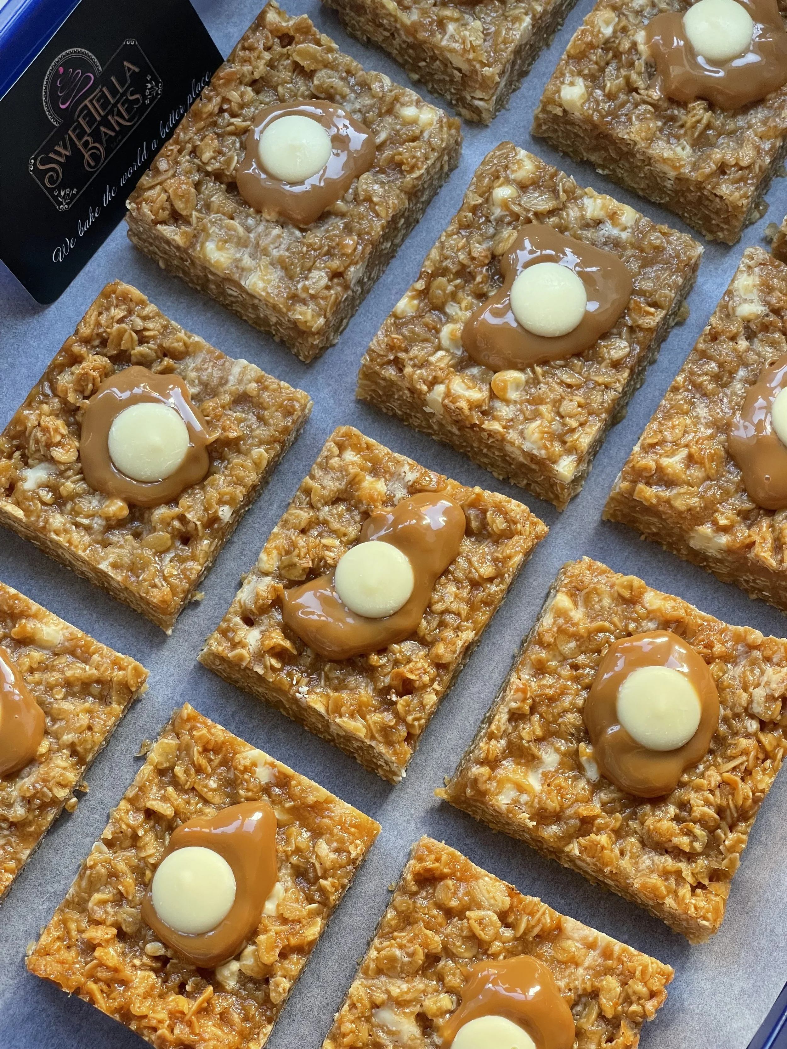 Close-up of caramel oat bars topped with white and caramel-colored chocolate discs, arranged on parchment paper.