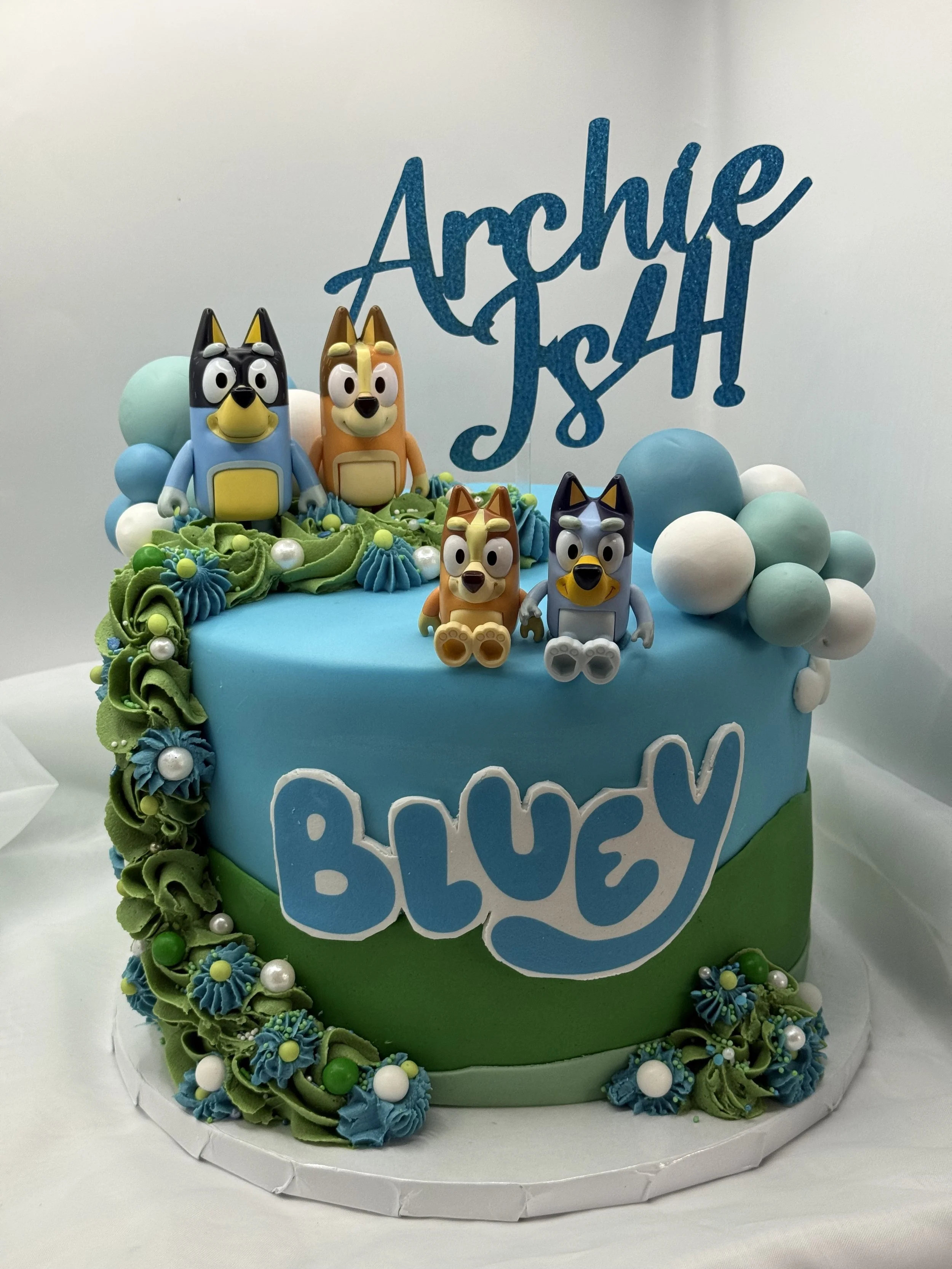 A birthday cake decorated with Bluey characters, colorful frosting, balloons, and a custom cake topper reading 'Archie Bash!'.