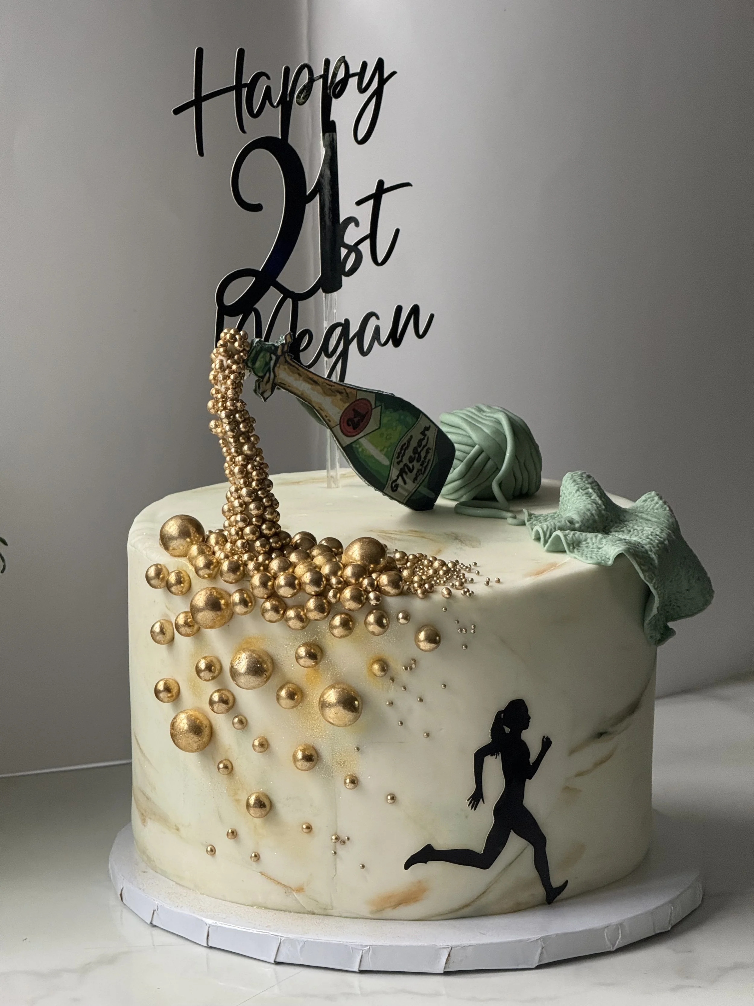 A celebration cake with a "Happy 21st Vegan" topper, gold beads, a running woman silhouette, a small green gift box, and a paper cutout of a champagne bottle, on a white base.