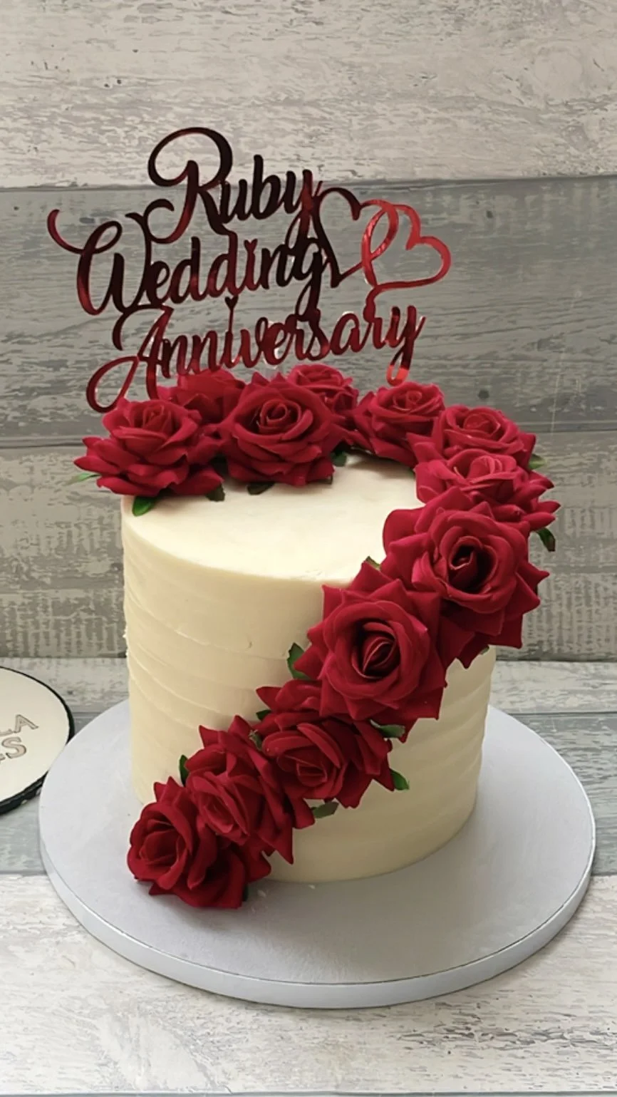 White wedding cake decorated with red roses and a topper reading "Ruby Wedding Anniversary" with a heart.
